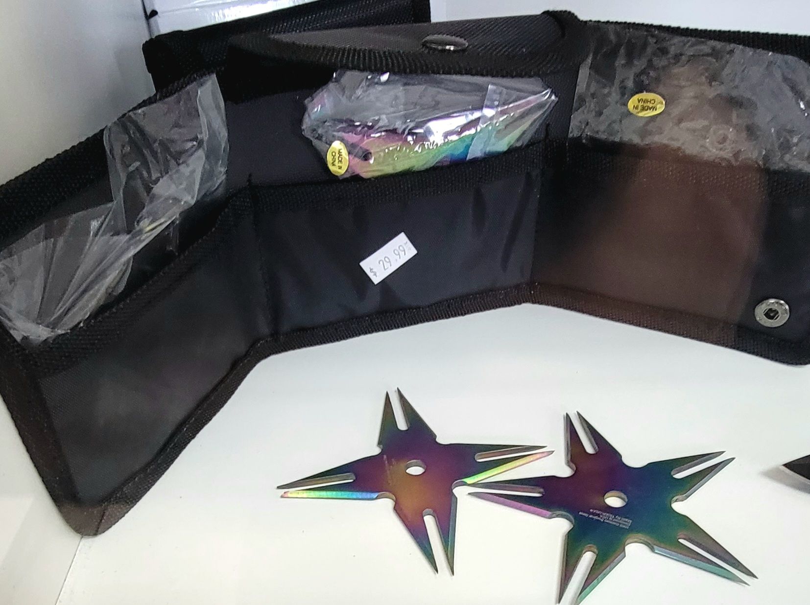 A black carrying case open on a white surface, showing rainbow-colored throwing stars and a packaged object.