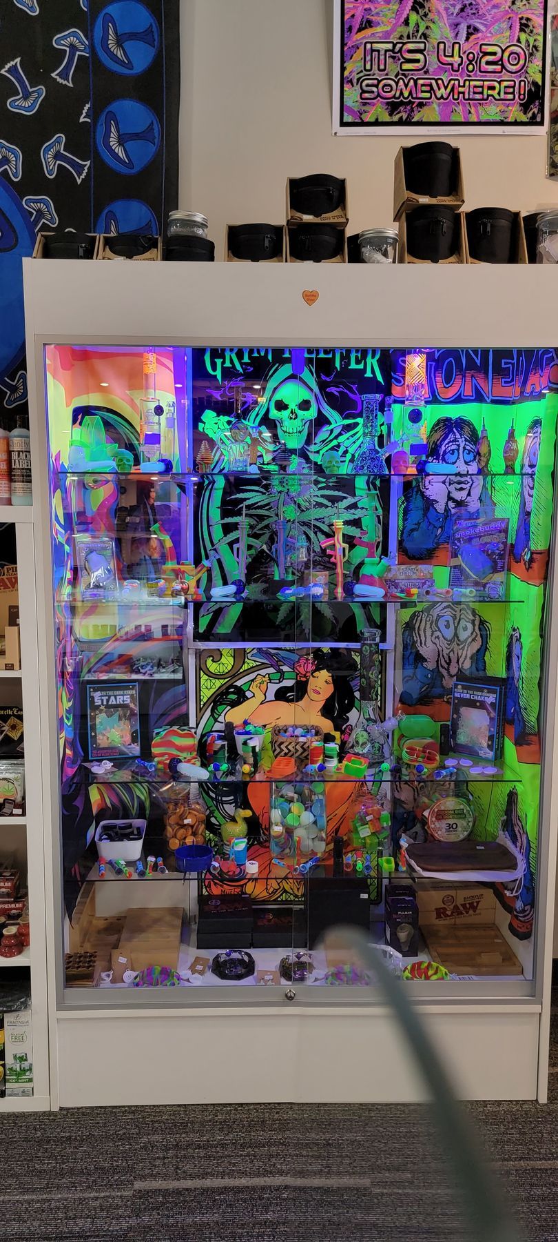 A white display cabinet illuminated with blacklight, filled with toys and art. A large green skeleton poster dominates the center.