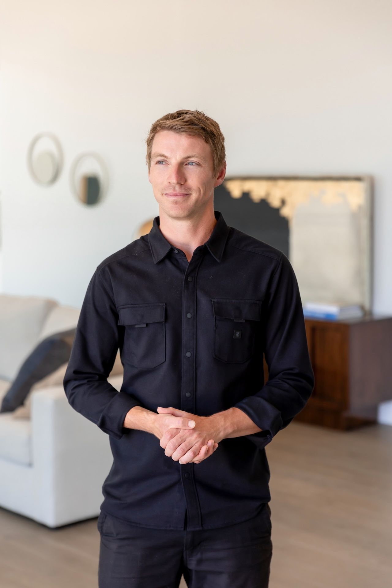 A man in a black shirt is standing in a living room with his hands folded.