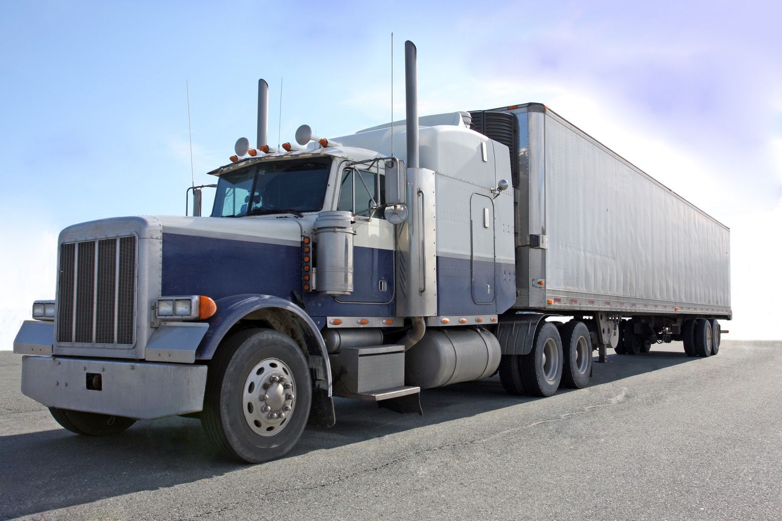 Blue and white semi-truck with a cargo trailer on a gravel road, under a light blue sky.