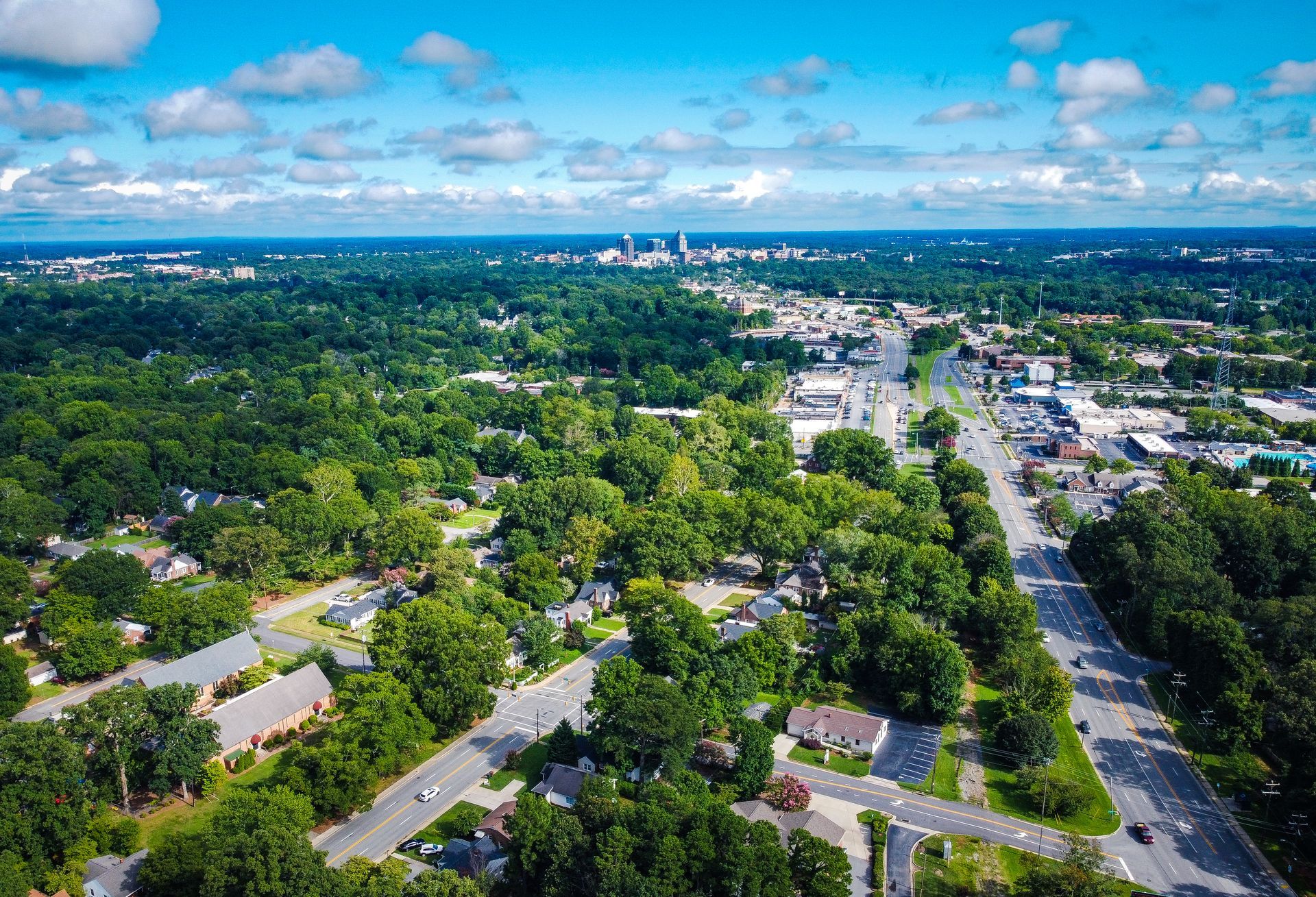 Aerial view of a residential Fort Mill SC neighborhood with dense trees leading to a distant city skyline under a blue, cloudy sky.