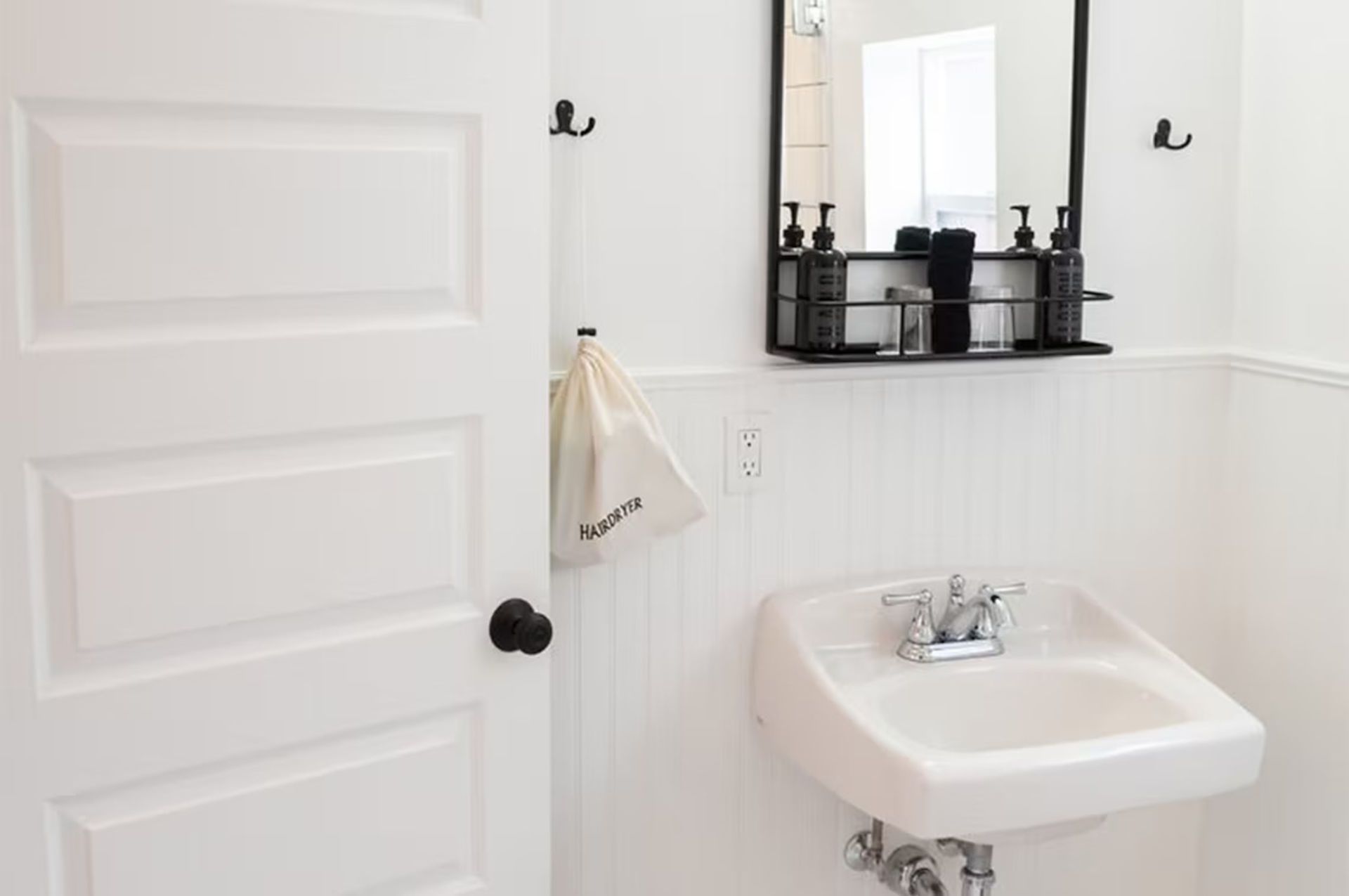 White bathroom with door ajar, sink, mirror, and black storage shelf.
