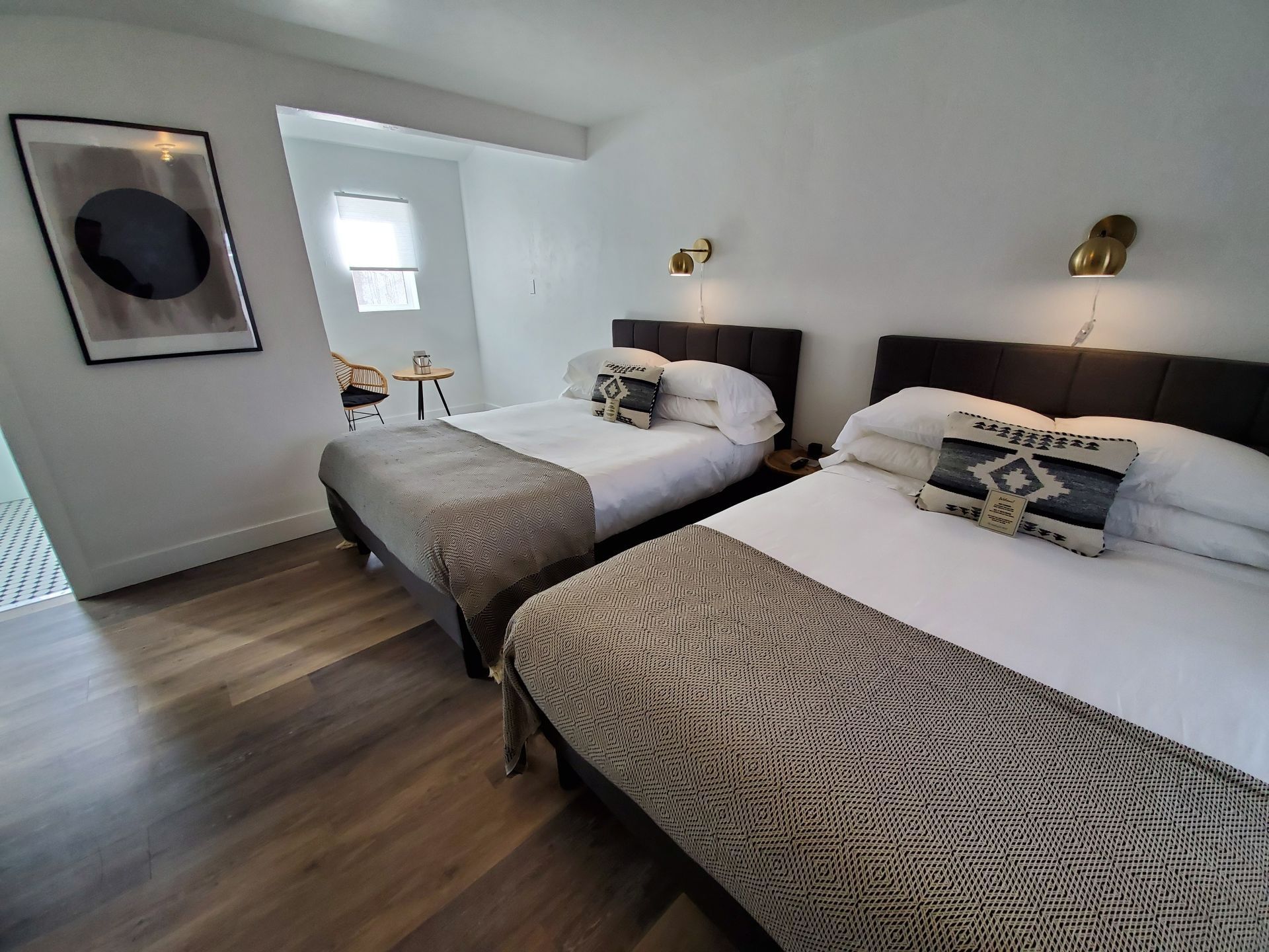 Two beds in a bright room, each with decorative pillows and patterned throw, artwork on the wall.