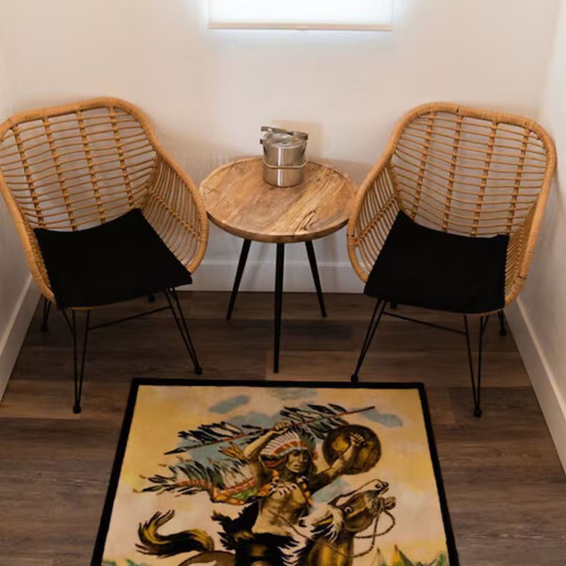 Two wicker chairs with black cushions, small round table, rug with cowboy artwork, under a window.