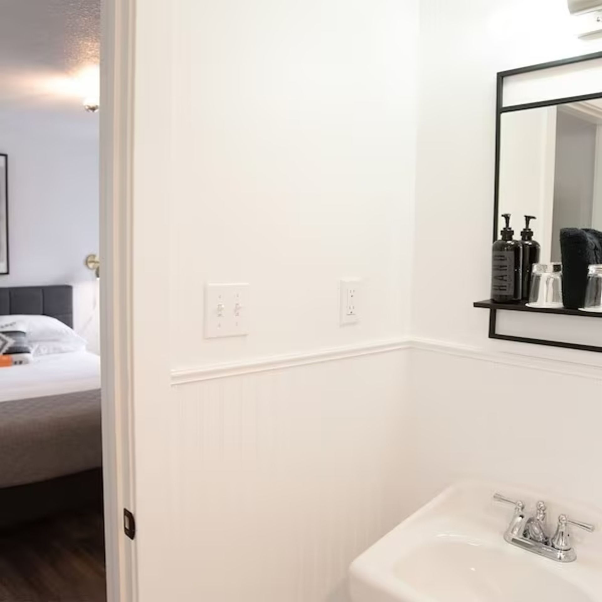 Bathroom interior with white walls, sink, mirror, and partially visible bedroom through doorway.