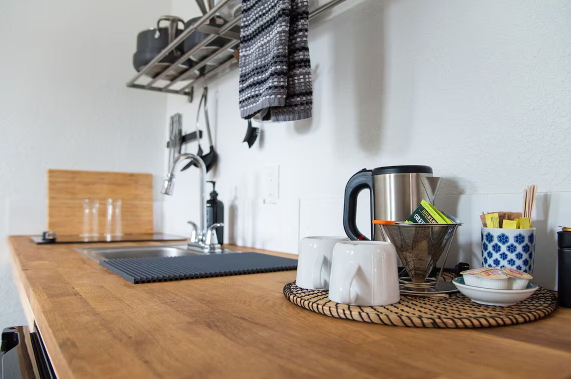 Kitchen counter with sink, kettle, cups, and various items on a wooden surface against a white wall.