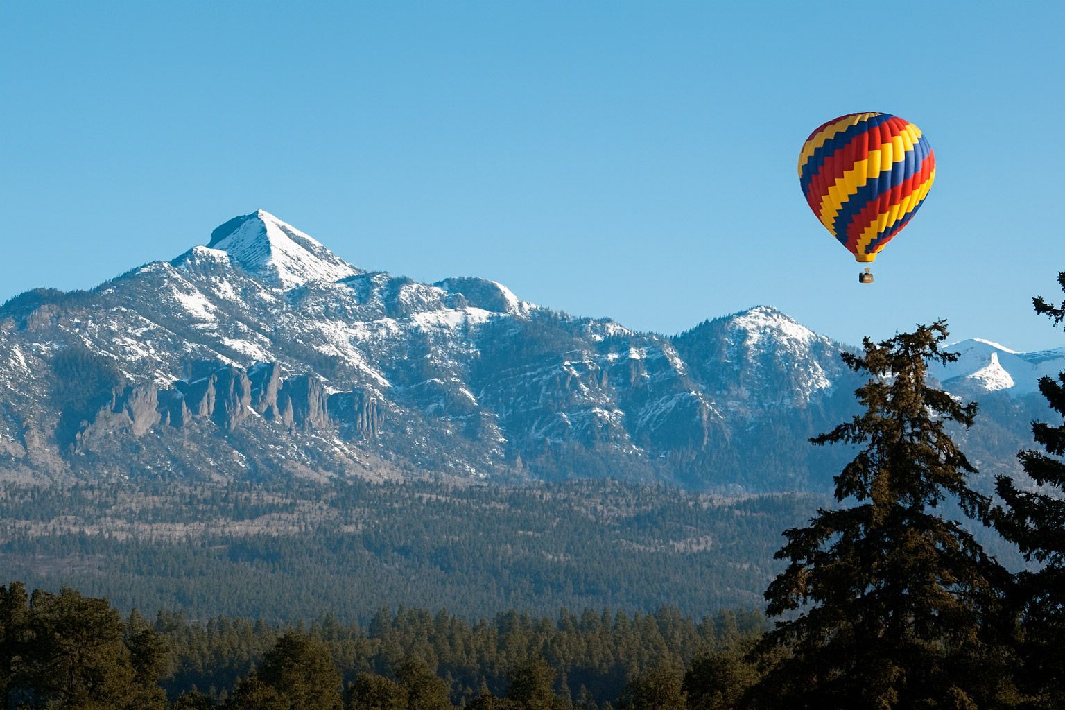 Hot air balloon soaring over snow-capped mountains and a forest.