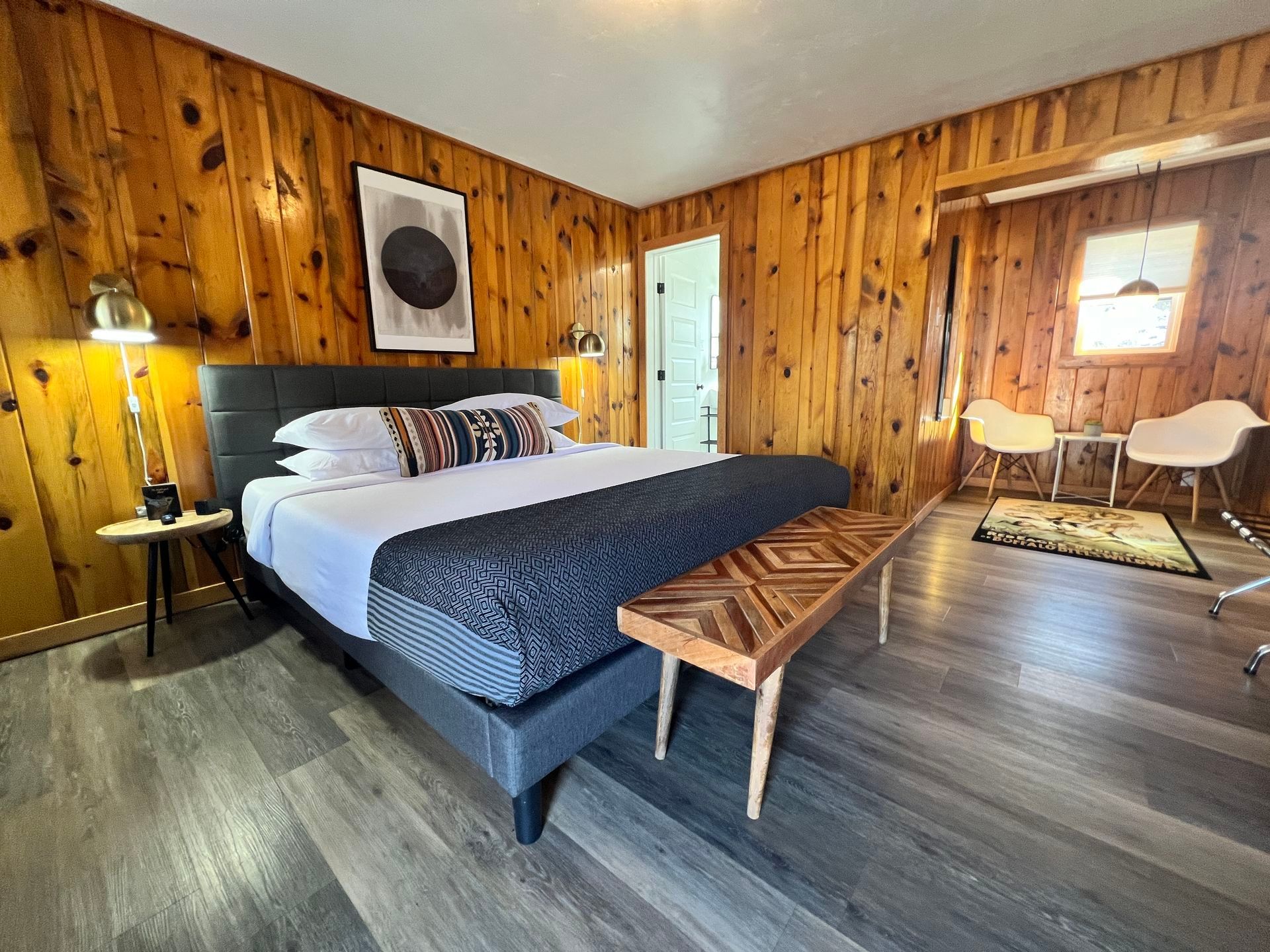 Bedroom with wood-paneled walls, a bed with decorative pillows, a bench, and two chairs near a window.