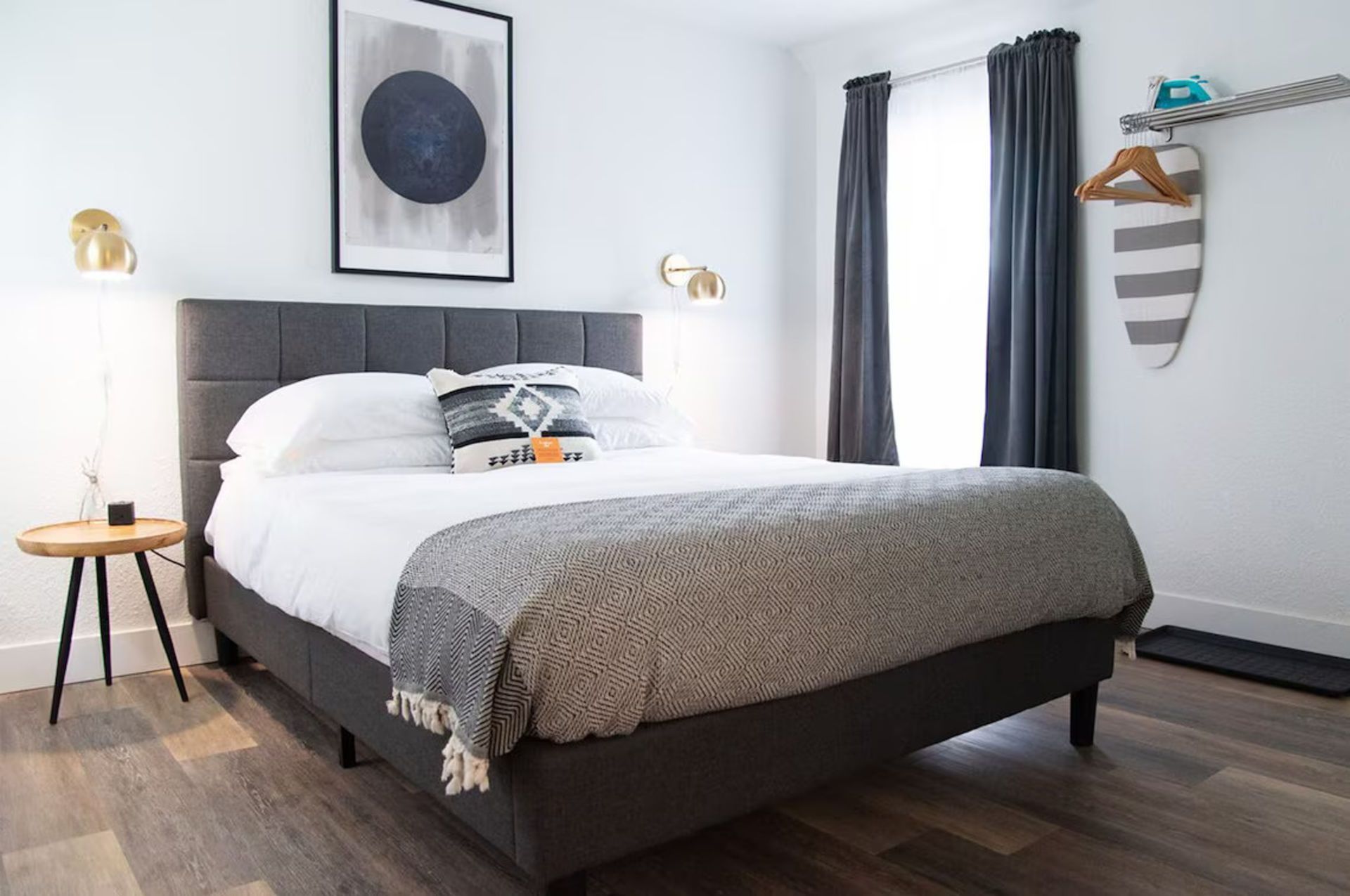 Bedroom with gray bed, art, bedside table, curtains, and ironing board.