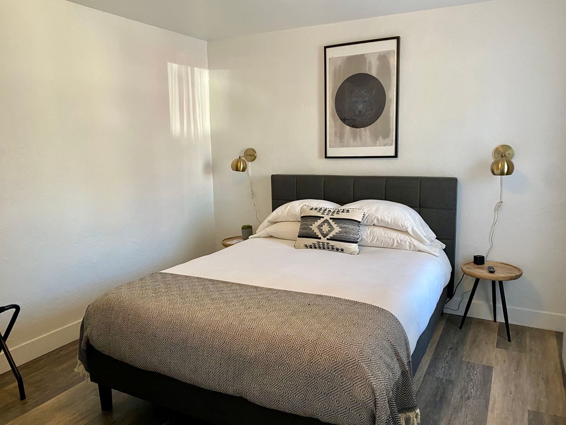 Bedroom with a bed, nightstands, art, and sconces. White walls, wood floor, and patterned blanket.