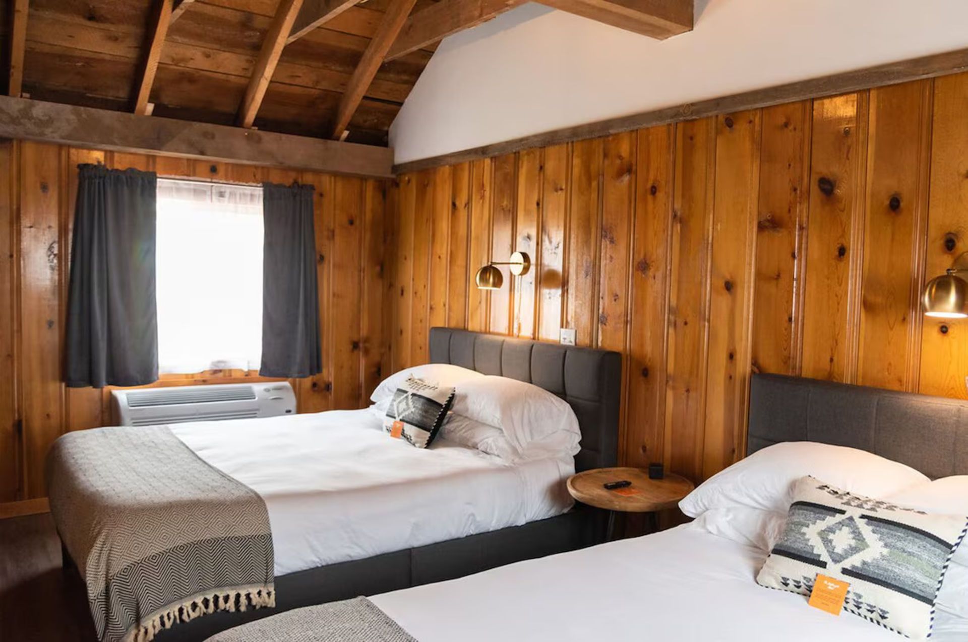 Two twin beds in a rustic room with wood paneling, window with gray curtains, and sconce lighting.