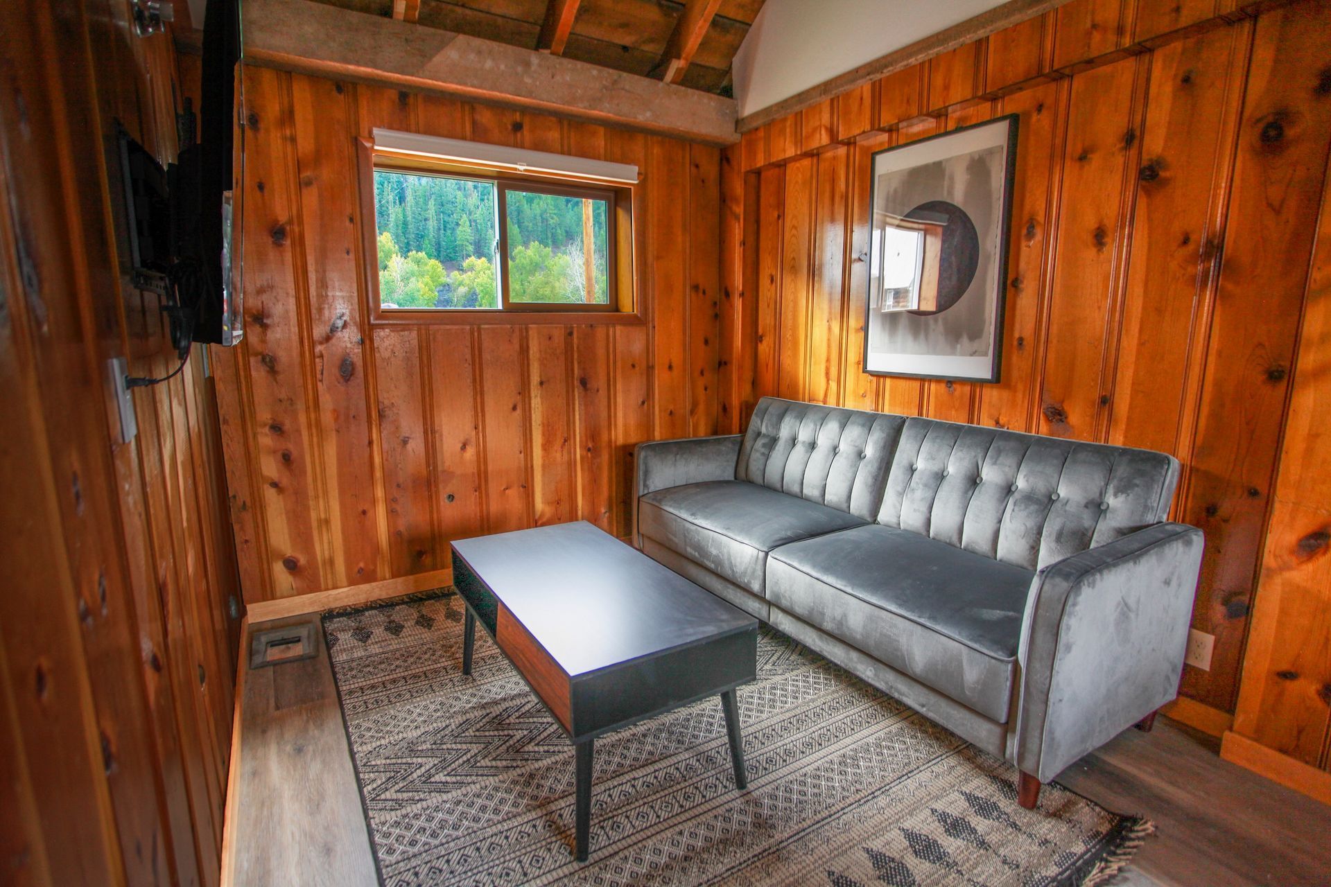 Cozy cabin living room with wood paneling, gray sofa, rug, coffee table, and window with forest view.