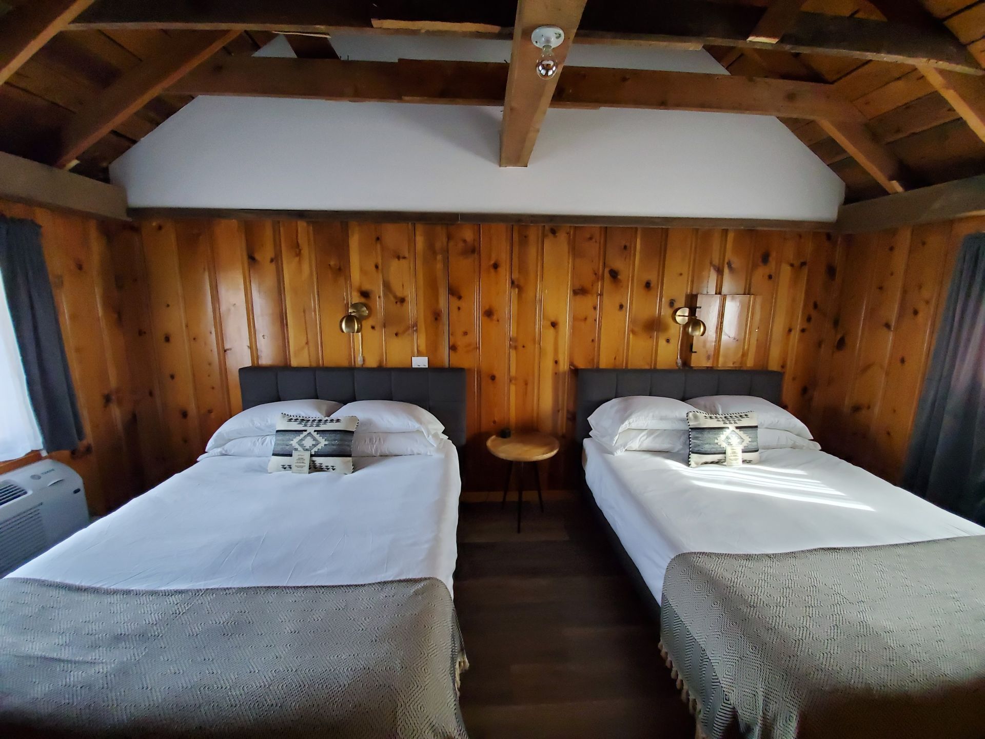Two twin beds in a wood-paneled room. Soft light shines on white sheets and gray blankets.
