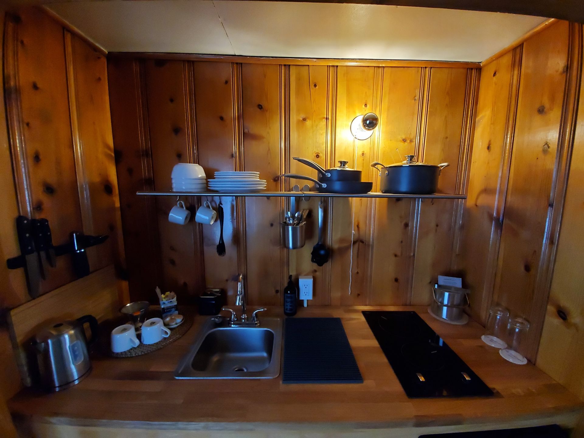 Small kitchen with wood paneling, a sink, stovetop, and shelves holding dishes and cookware.