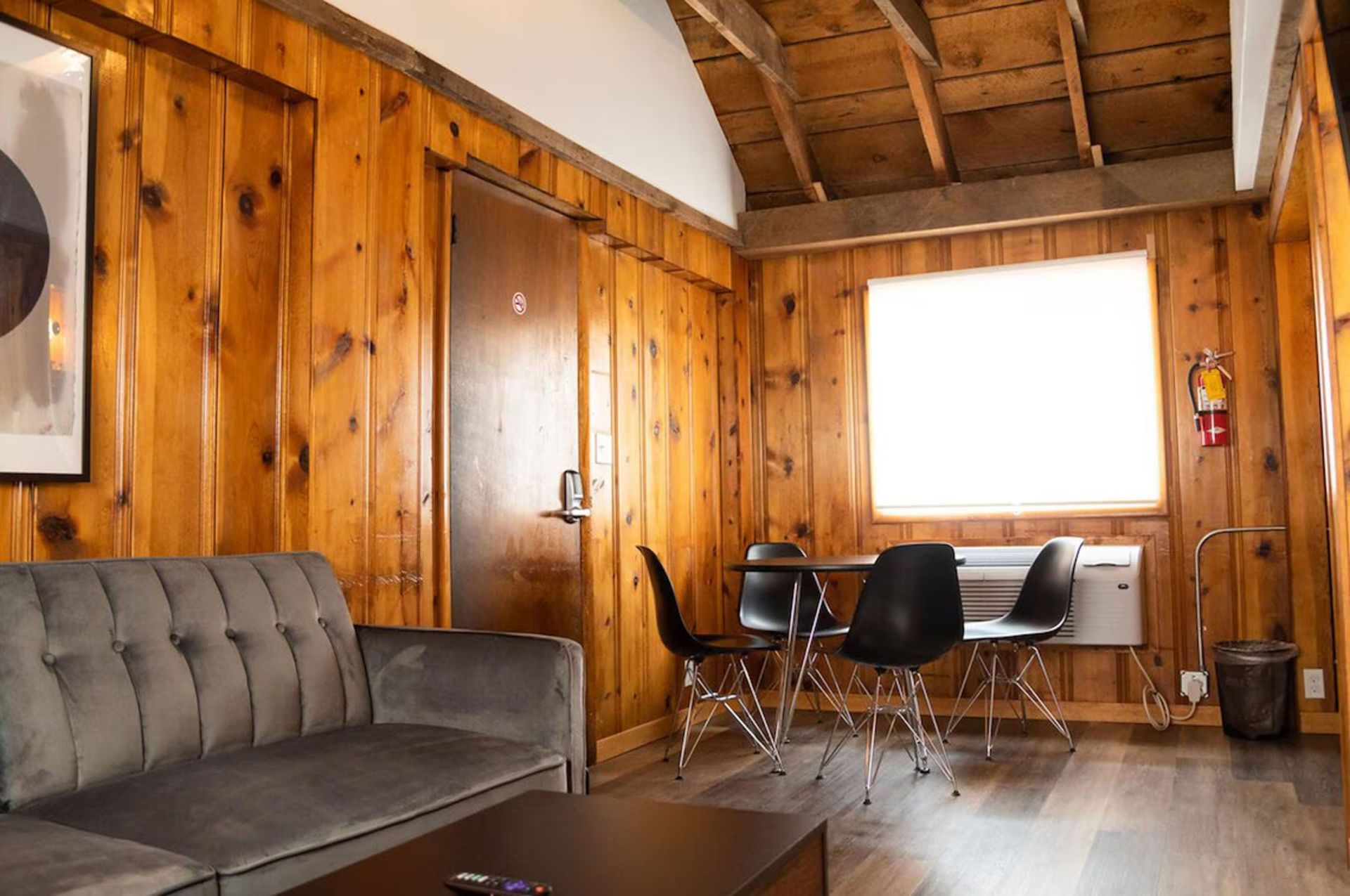 Cozy cabin interior with wood paneling, gray sofa, black table & chairs, a window, and a fire extinguisher.