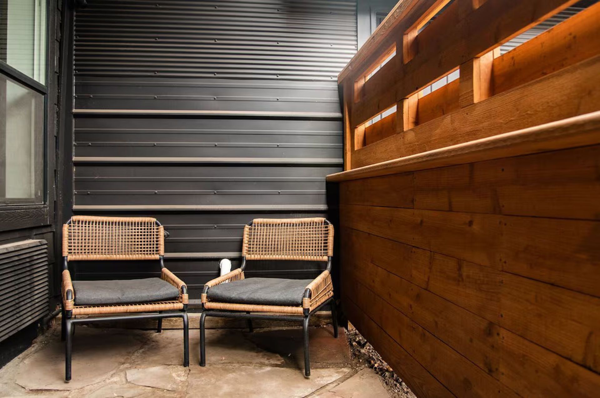 Two wicker chairs with gray cushions sit on a stone patio, near a dark metal wall and wooden fence.