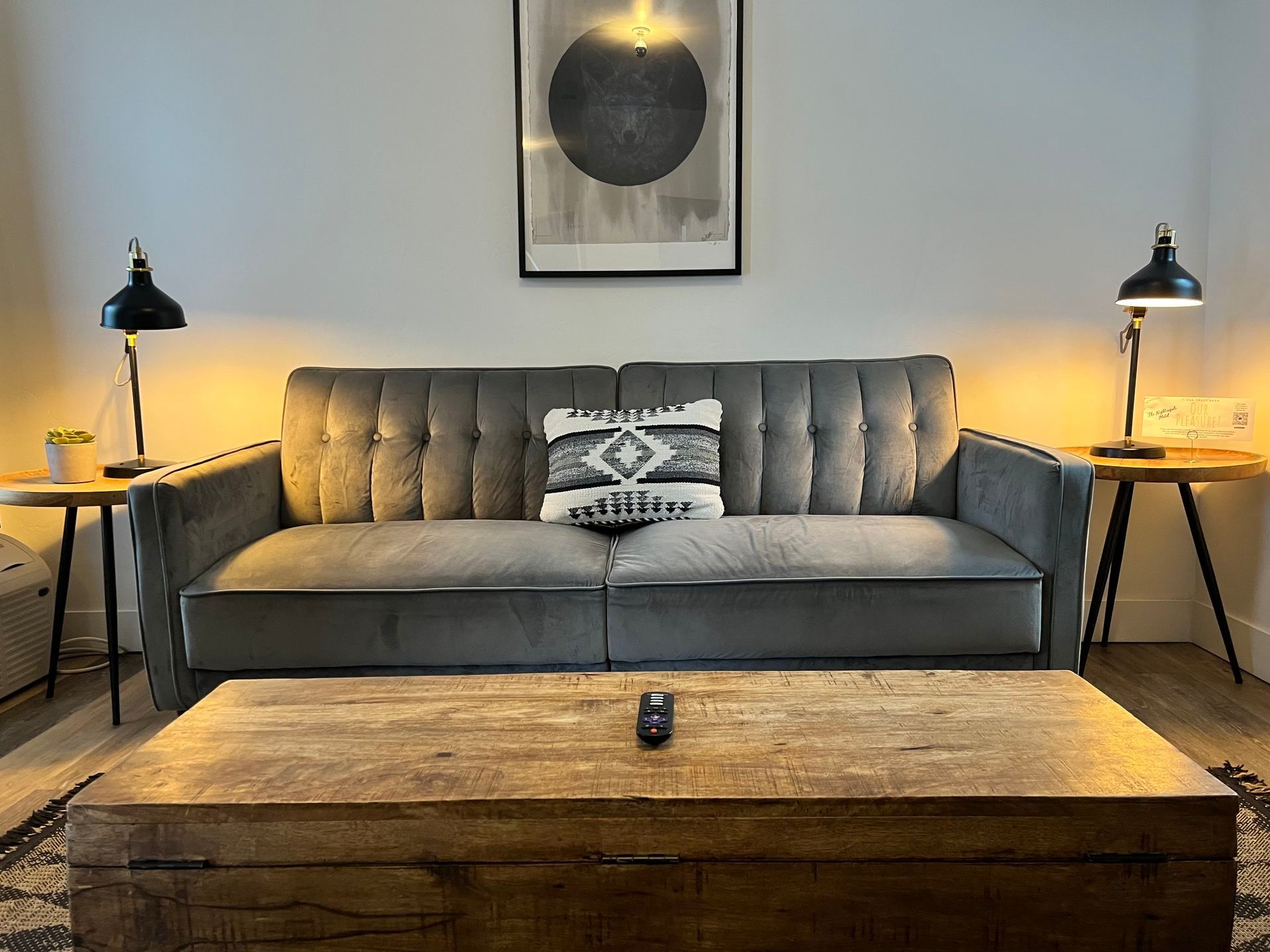 Gray couch with a throw pillow between two black lamps on end tables, and a rustic coffee table.