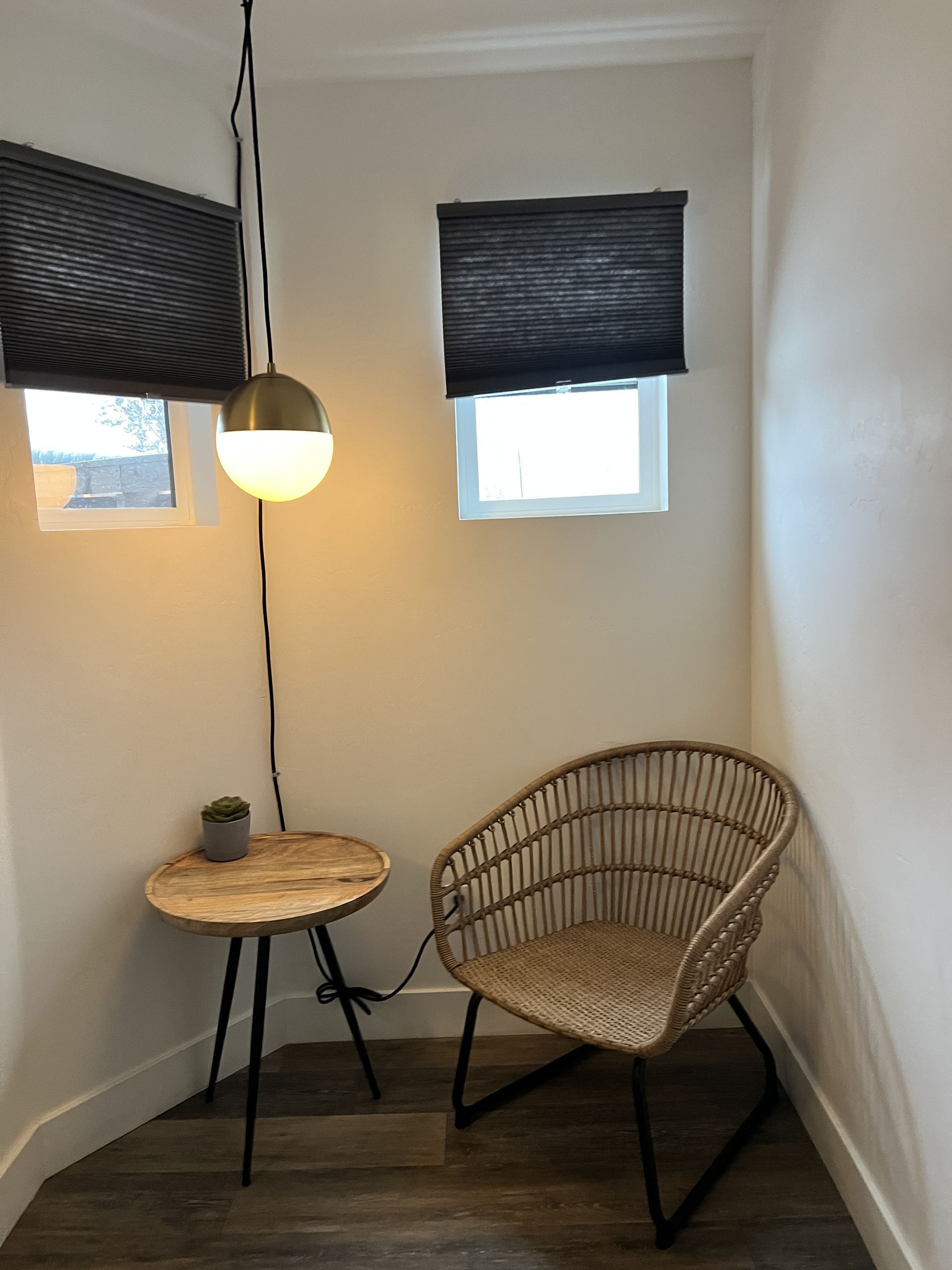 Cozy corner with wicker chair, small table, and hanging globe light. Two windows with dark shades.