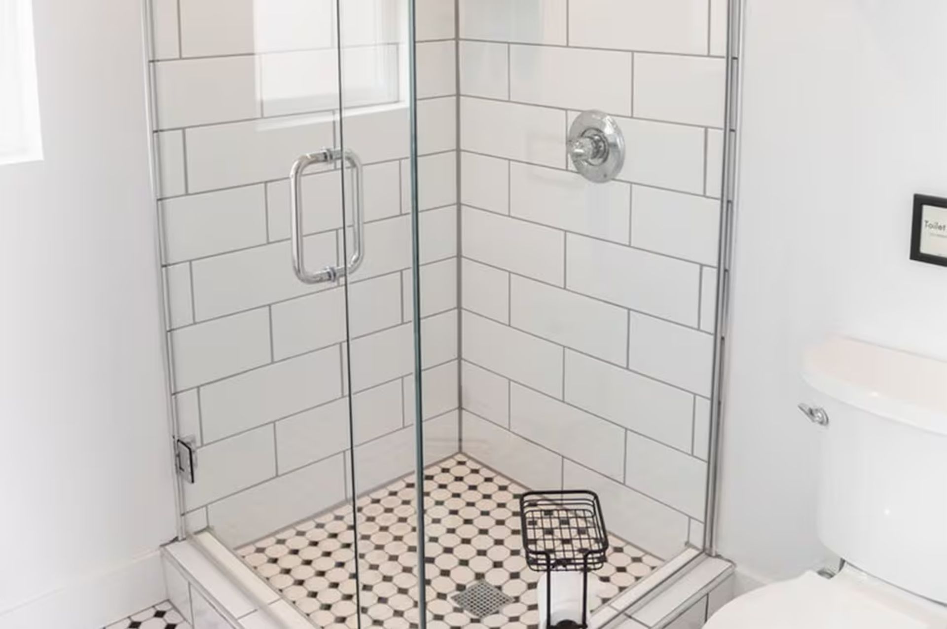 White-tiled shower with glass door, black and white tiled floor. Toilet to the right, metal basket inside.