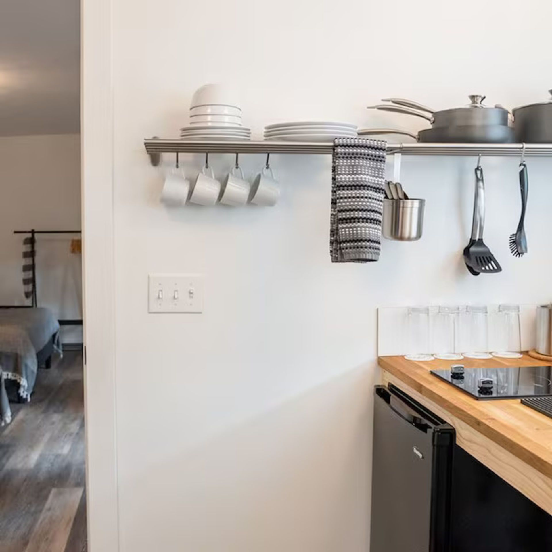 Small kitchen with stainless steel rack holding dishes, mugs, utensils, and a towel. Black stovetop and mini-fridge.