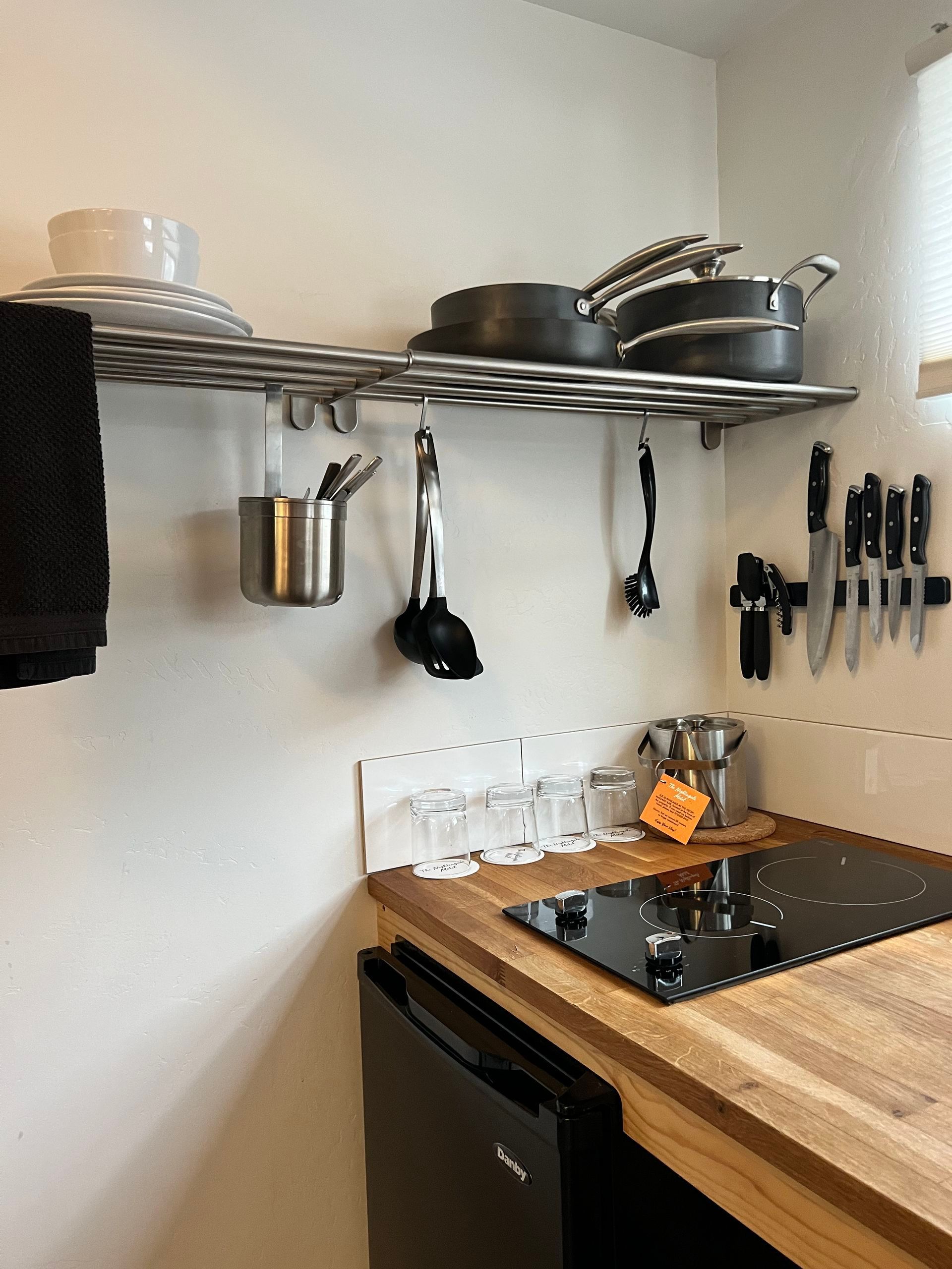 Small kitchen with pots, pans, utensils, and knives on a shelf above a stove. Black appliance.