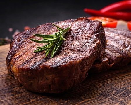 Two grilled steaks on a wooden board, garnished with rosemary, with red peppers in the background.