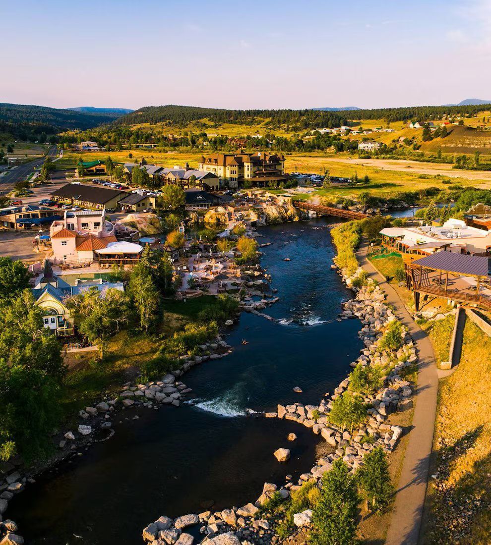 Aerial view of a town along a river, with buildings, green hills, and a walking path along the water.