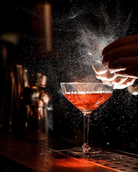 Bartender spritzing citrus oils over a red cocktail in a coupe glass at a dimly lit bar.