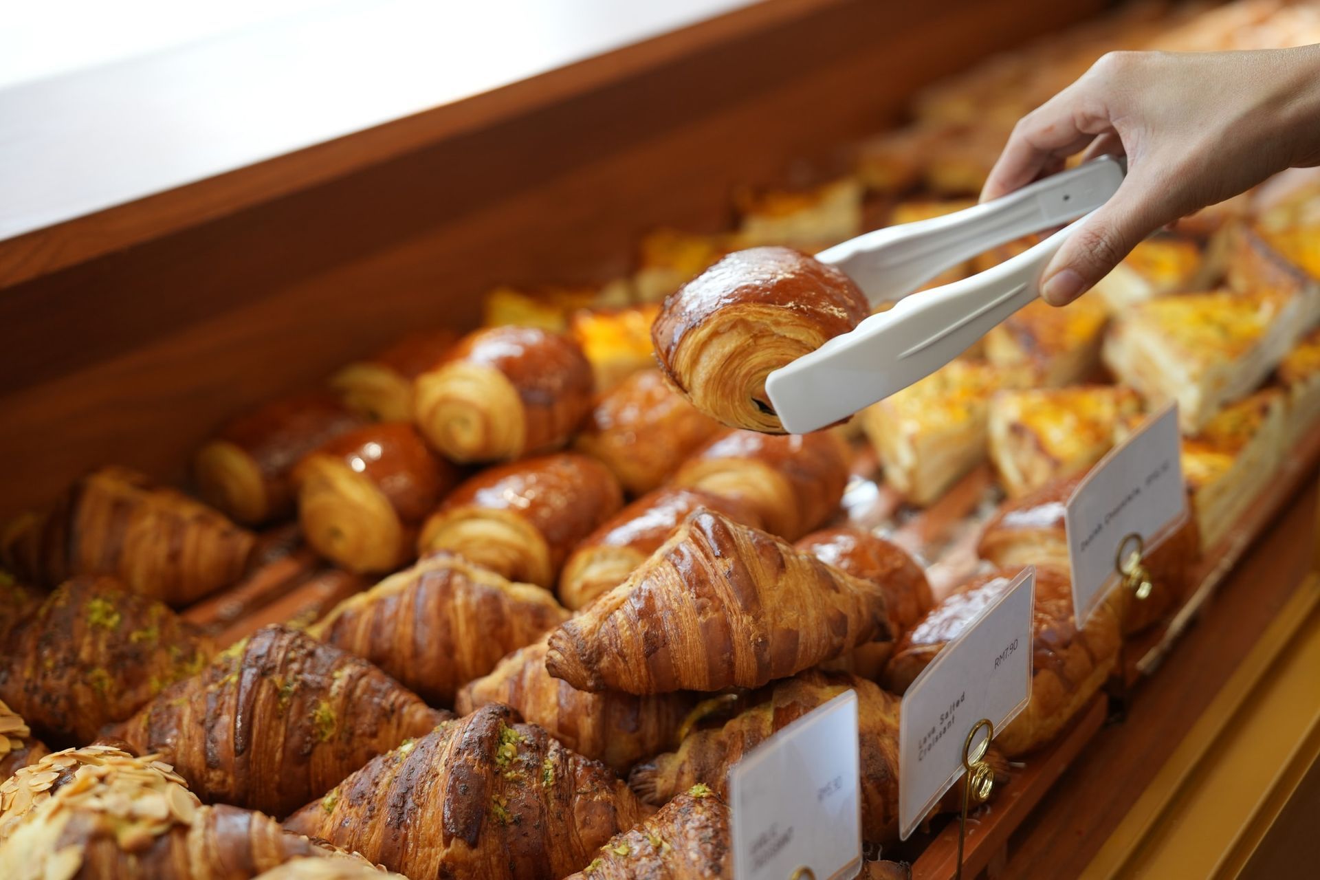 Hand using tongs to pick a croissant from a bakery display filled with pastries.