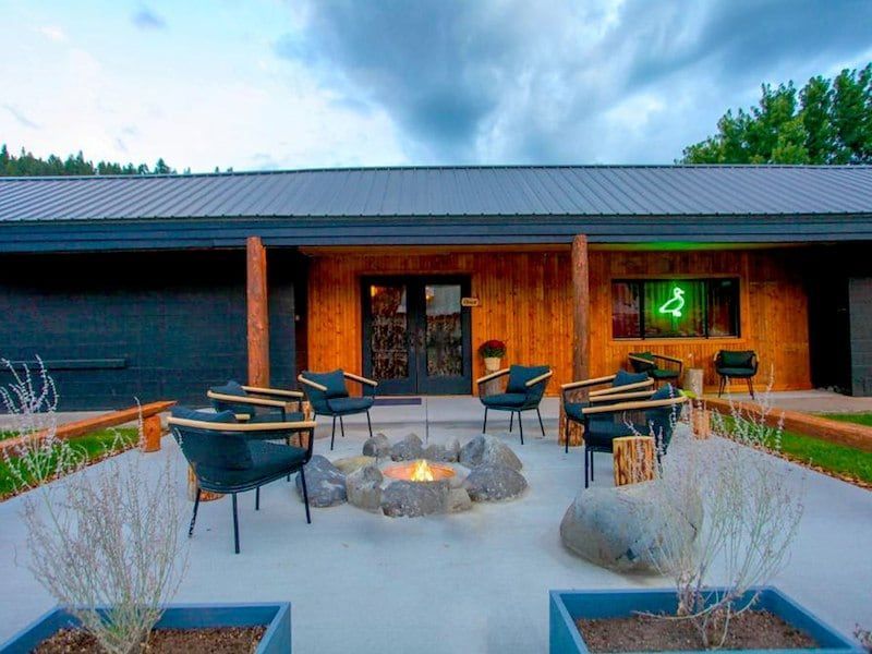 Exterior of a modern building with a fire pit and chairs on a patio. Green neon sign, gray cloudy sky.