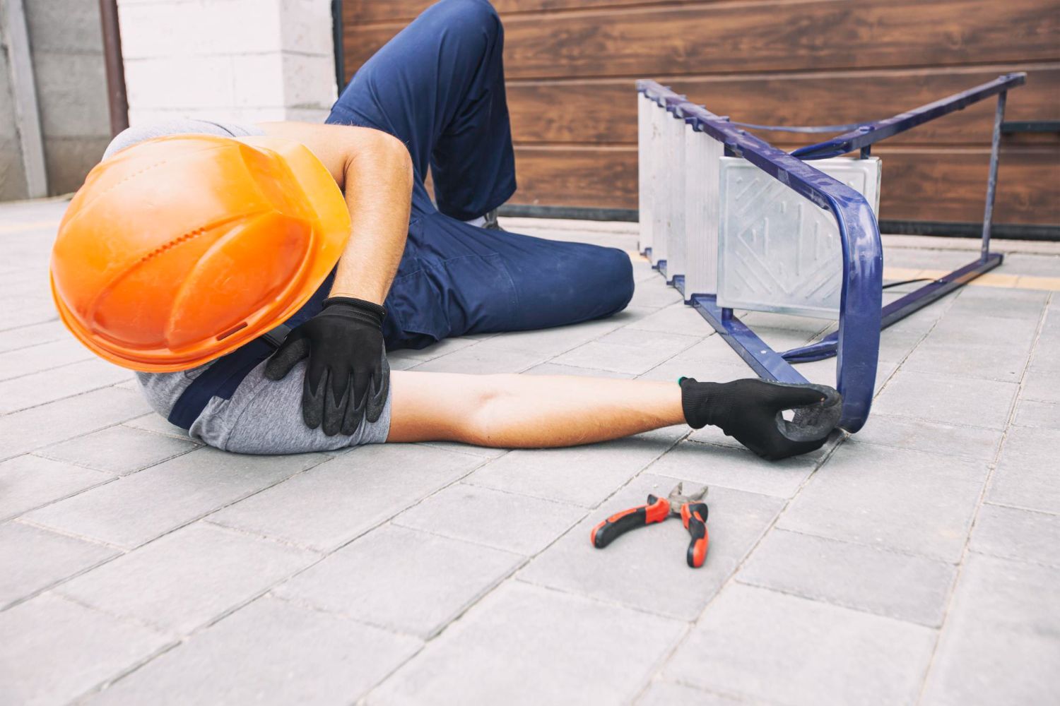 Worker in orange hard hat lying on pavement beside a damaged blue hand truck and tools.