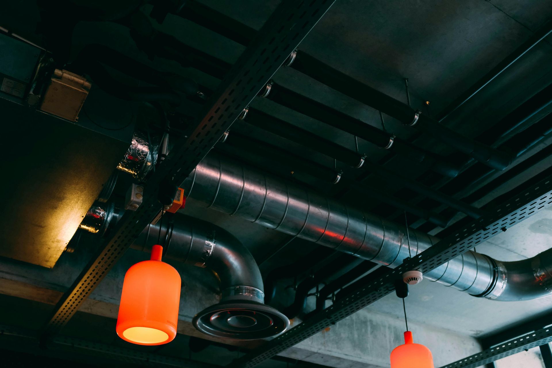Industrial ceiling with exposed pipes, ducts, and orange pendant lights.