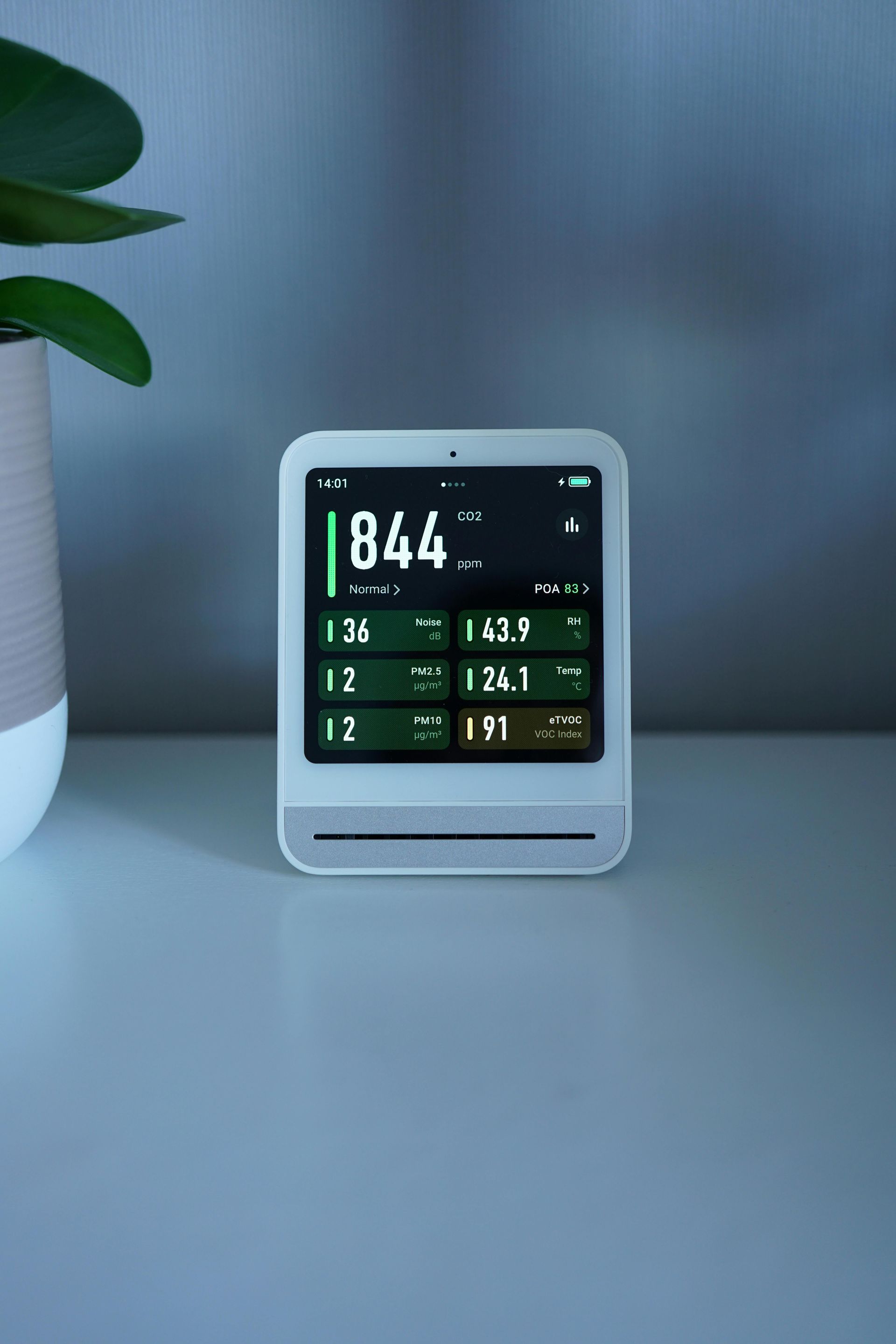 White air quality monitor displaying readings in green and white, on a white surface. A plant is beside it.