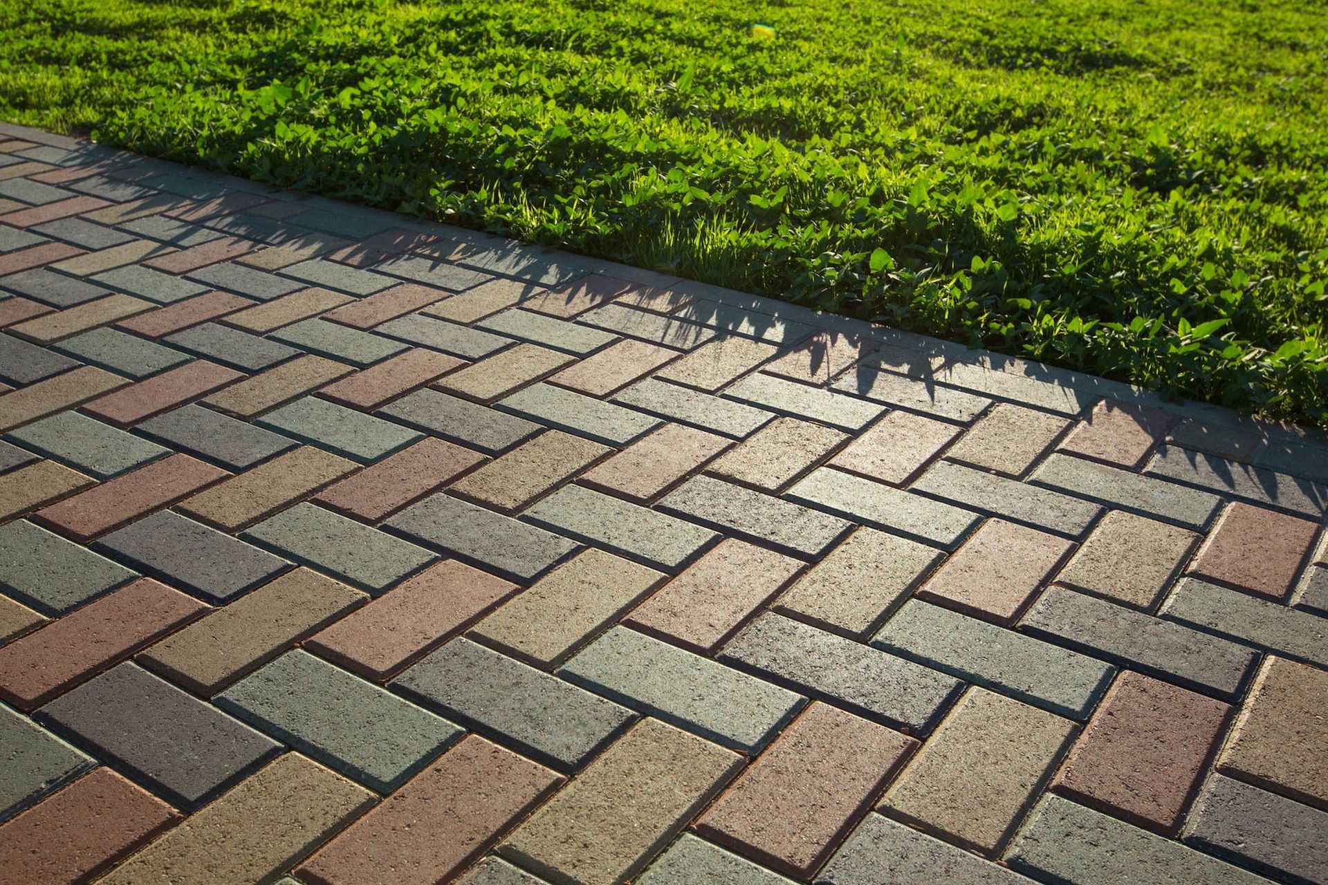 Herringbone pattern brick walkway in tan, gray, and red tones, bordering a patch of bright green grass.