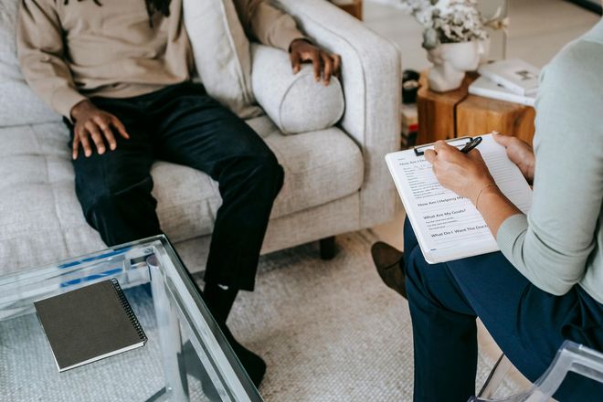 A man is sitting on a couch talking to a woman who is holding a clipboard.