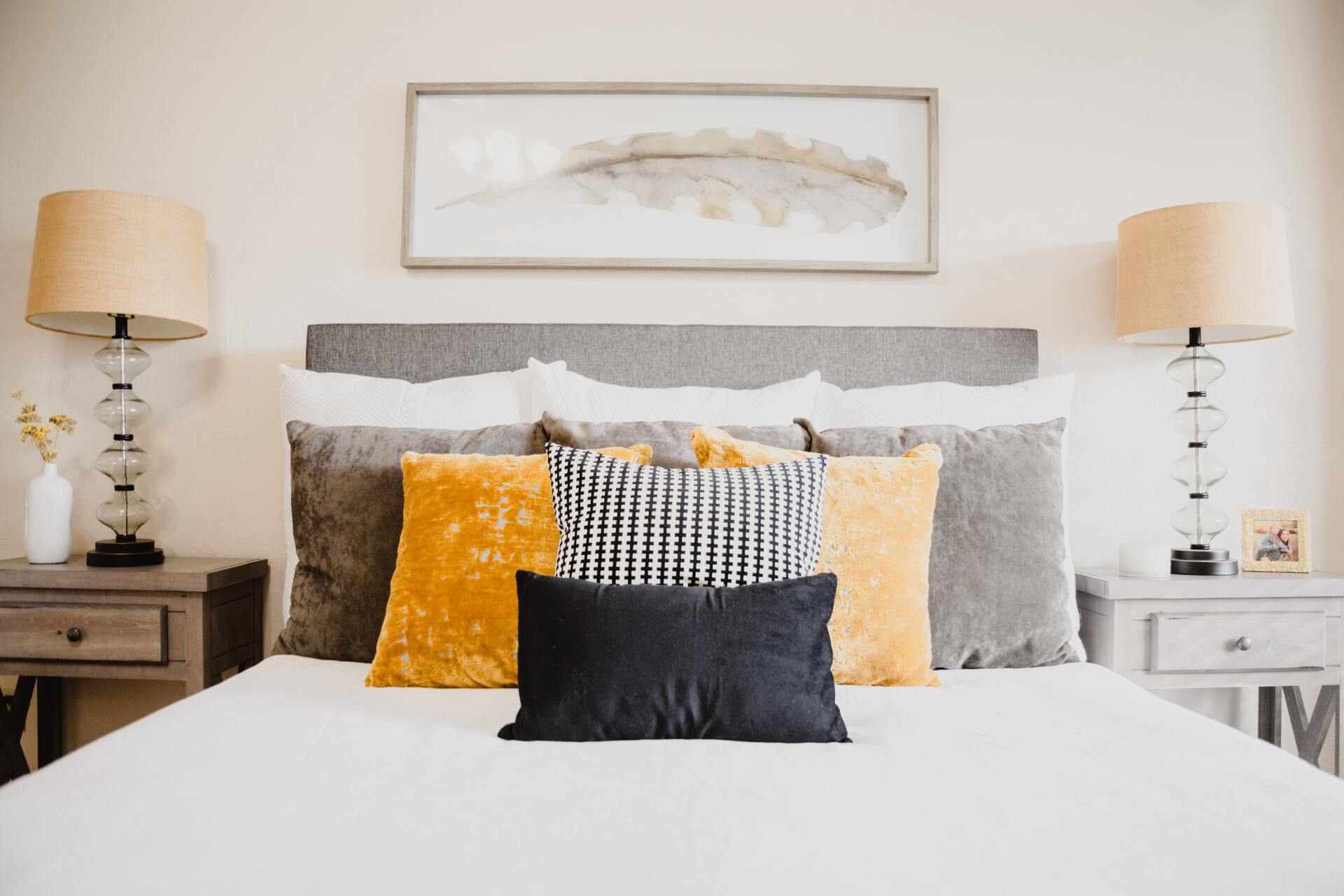 Bed with white sheets, a grey headboard, and colorful pillows on top. The bed is flanked with two lamps