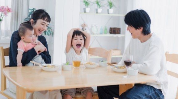 一家四口在餐桌旁一邊吃飯一邊歡笑。母親抱著嬰兒，父親面帶微笑。