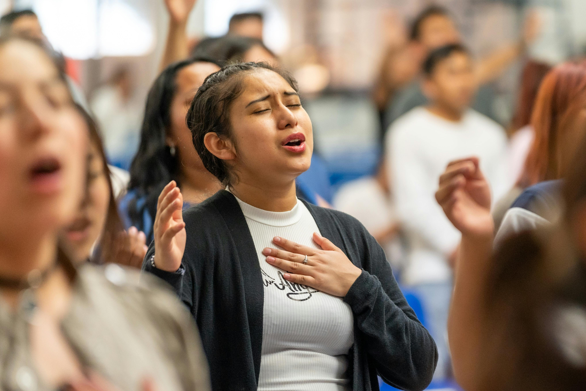 Group of people with arms raised, singing, some with eyes closed, in a brightly lit room.