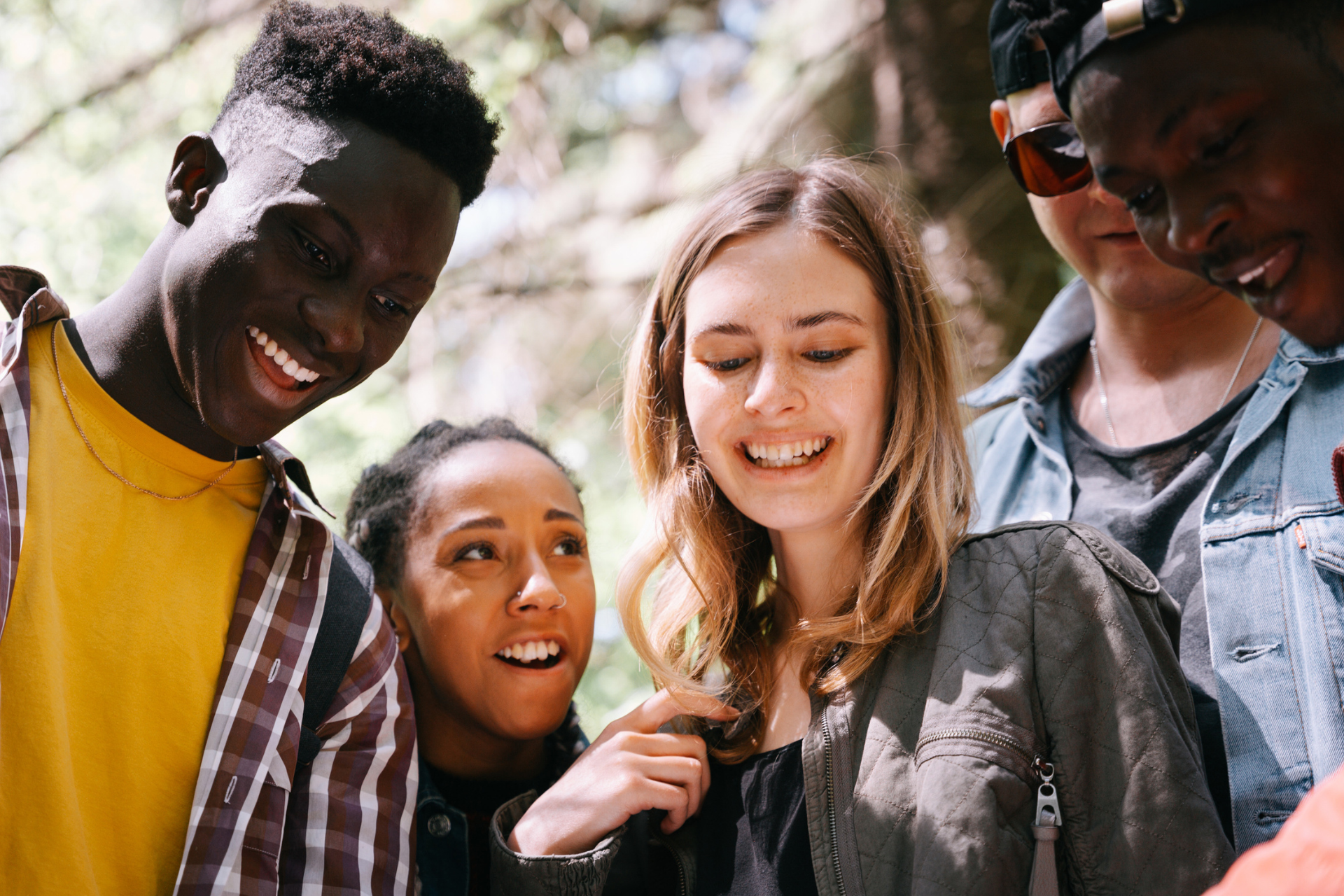 Group of smiling friends looking at something outdoors. Sunlight, trees, and jackets visible.