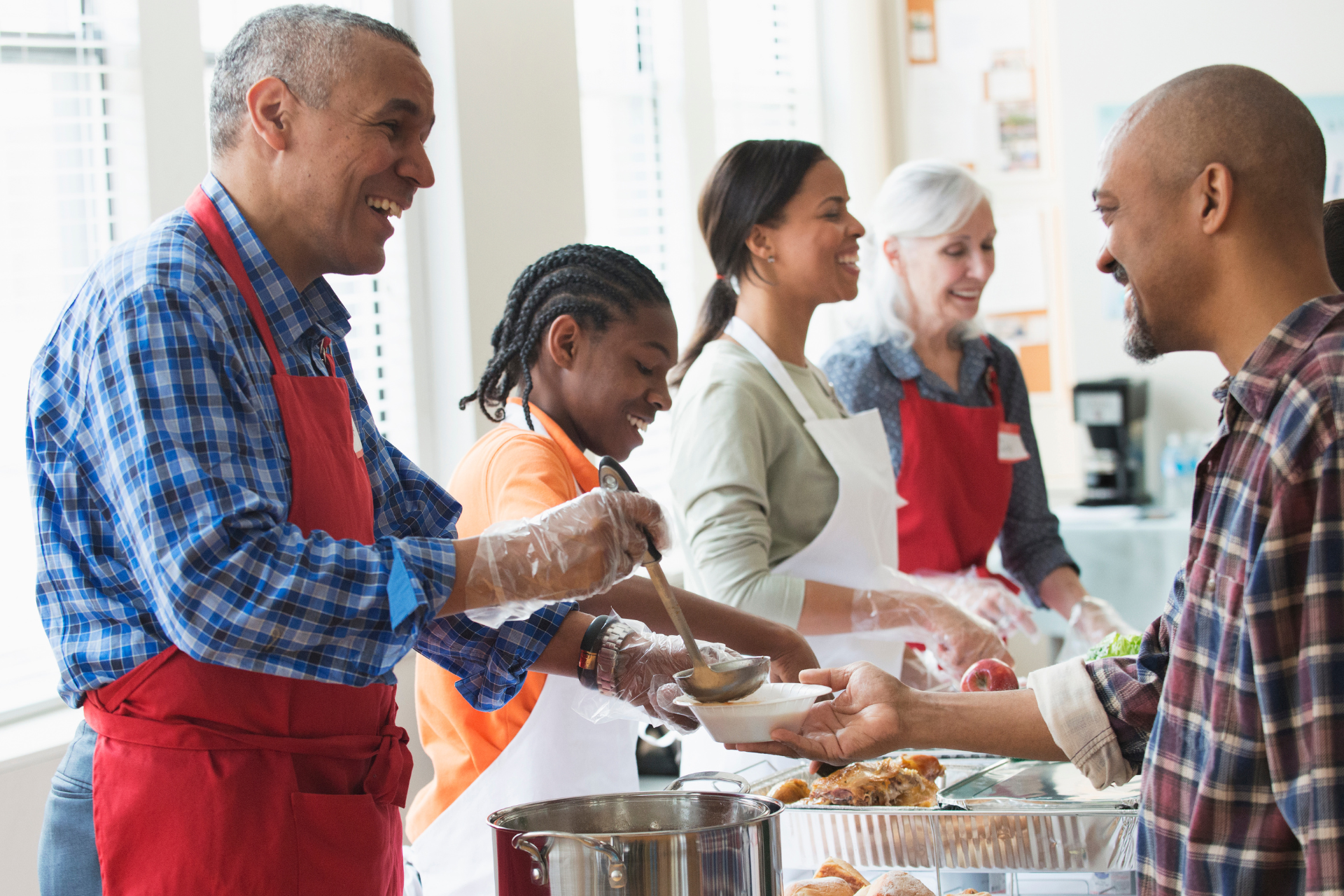 Volunteers serving food at a soup kitchen; smiling people, red aprons, white bowls, setting.