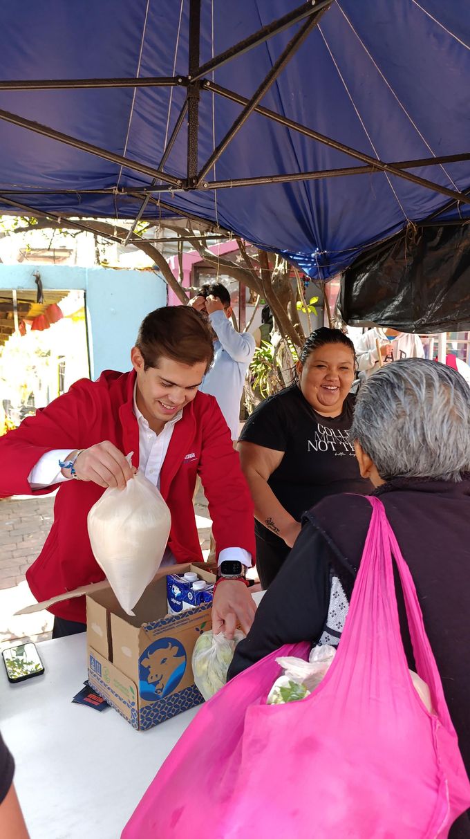 Man serving food under a blue canopy at a market stall, with people waiting nearby.