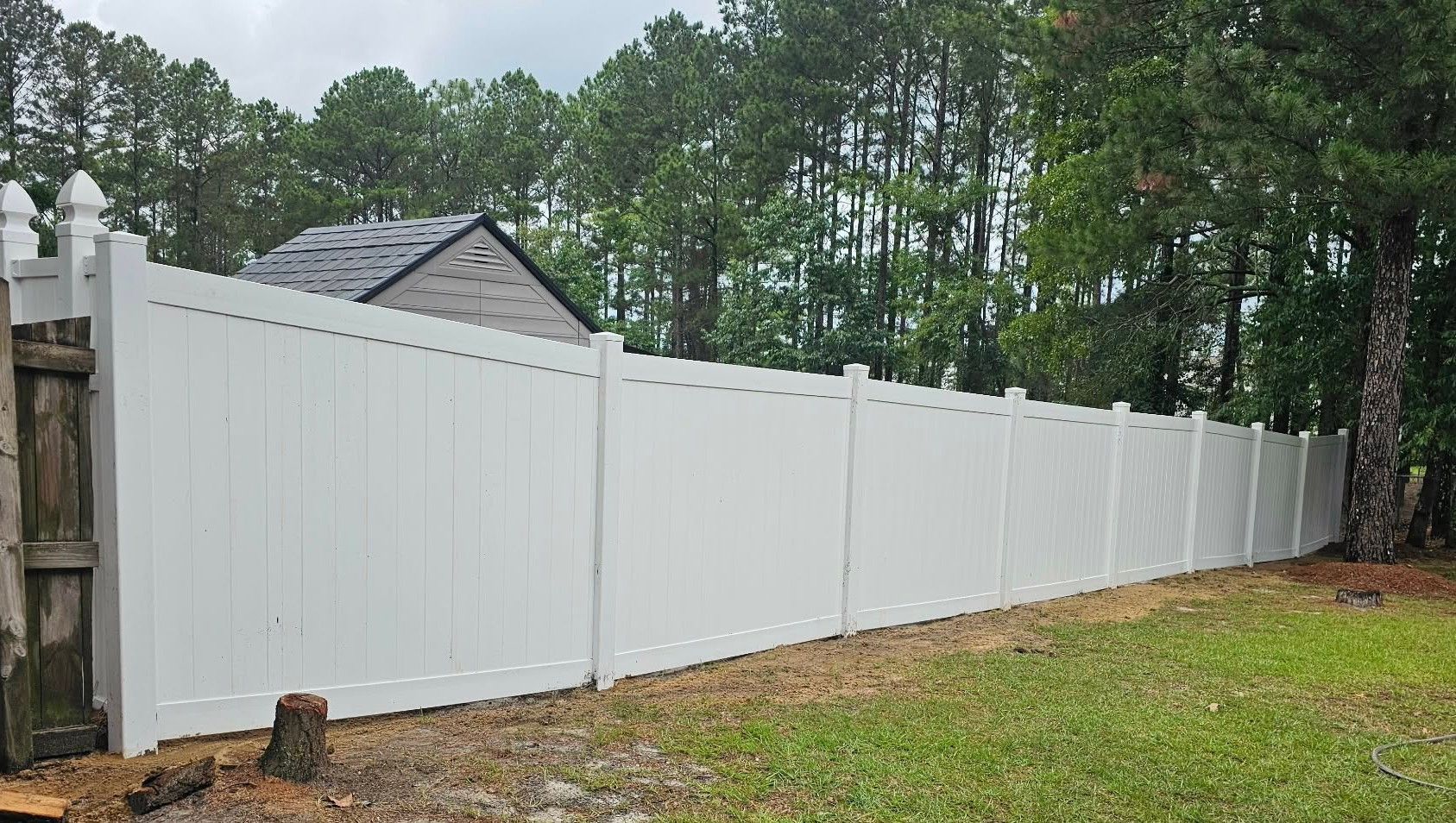 White vinyl privacy fence along a wooded backyard with a house roof visible behind it