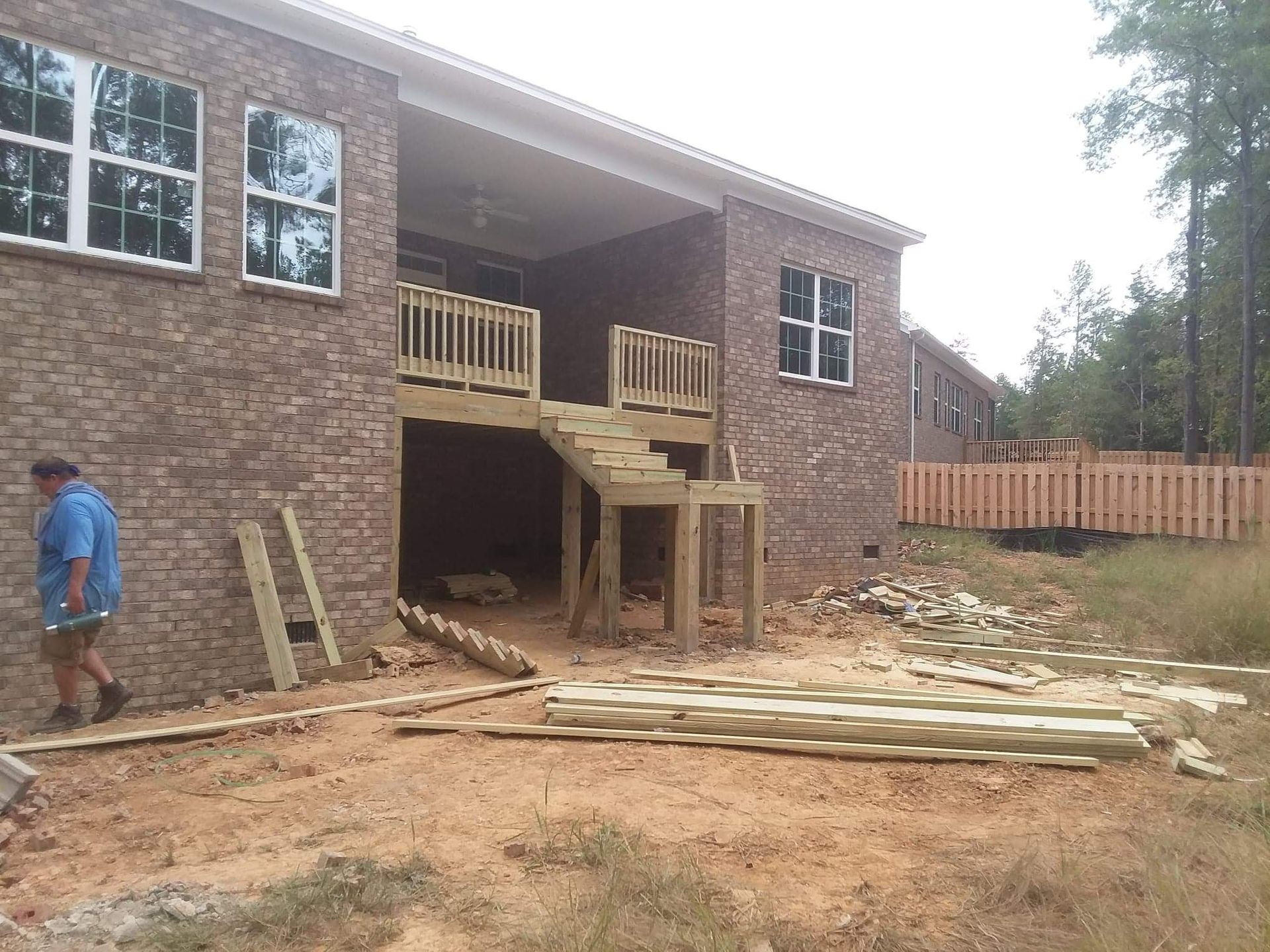 A man is standing in front of a brick house with a wooden deck under construction.