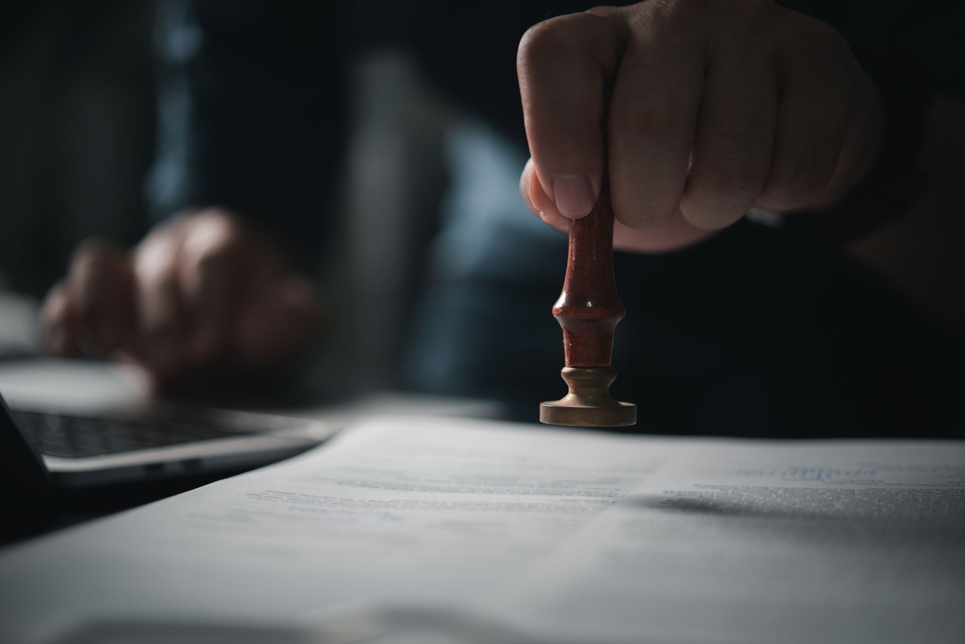 Hand holding a stamp, pressing it onto a white document on a desk, blurred background with laptop.