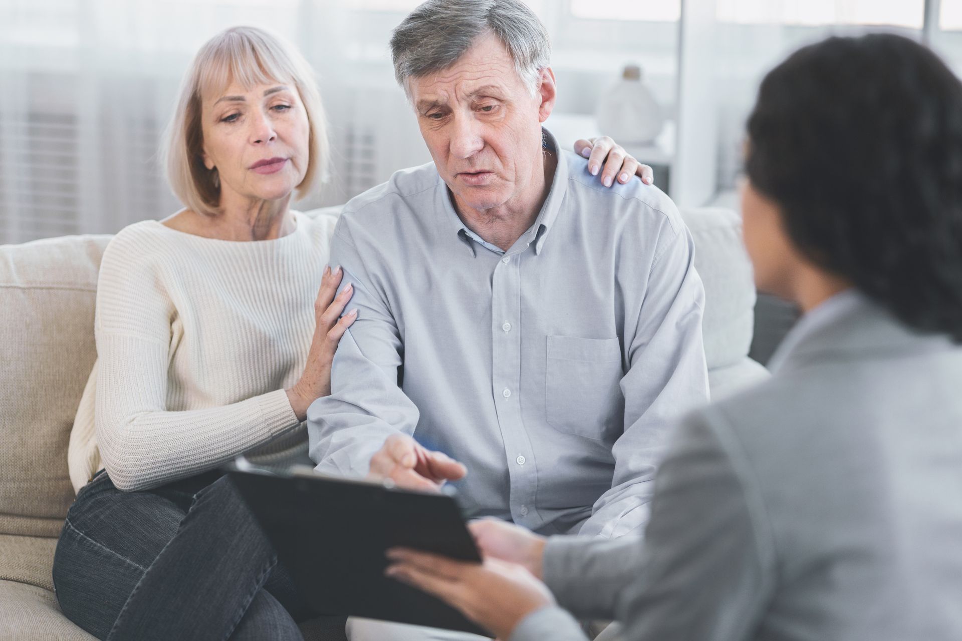 Senior couple consults with an advisor, discussing documents on a tablet; sitting indoors.