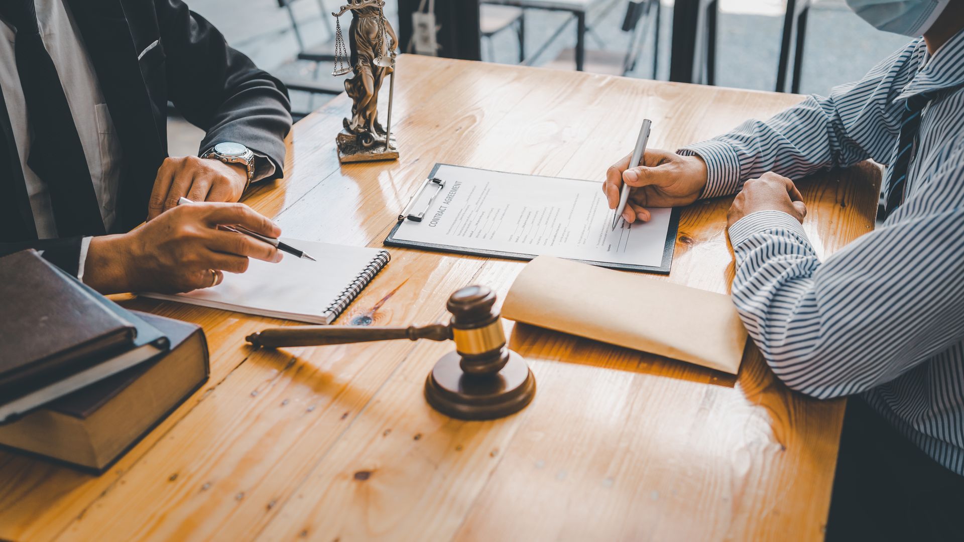Two people at a wooden table reviewing documents, one signing. Gavel, law books, and a small statue are also on the table.