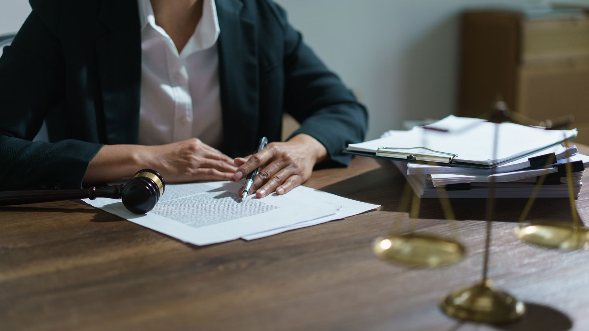Person in a blazer reviewing documents, with a gavel and scales of justice on a wooden desk.