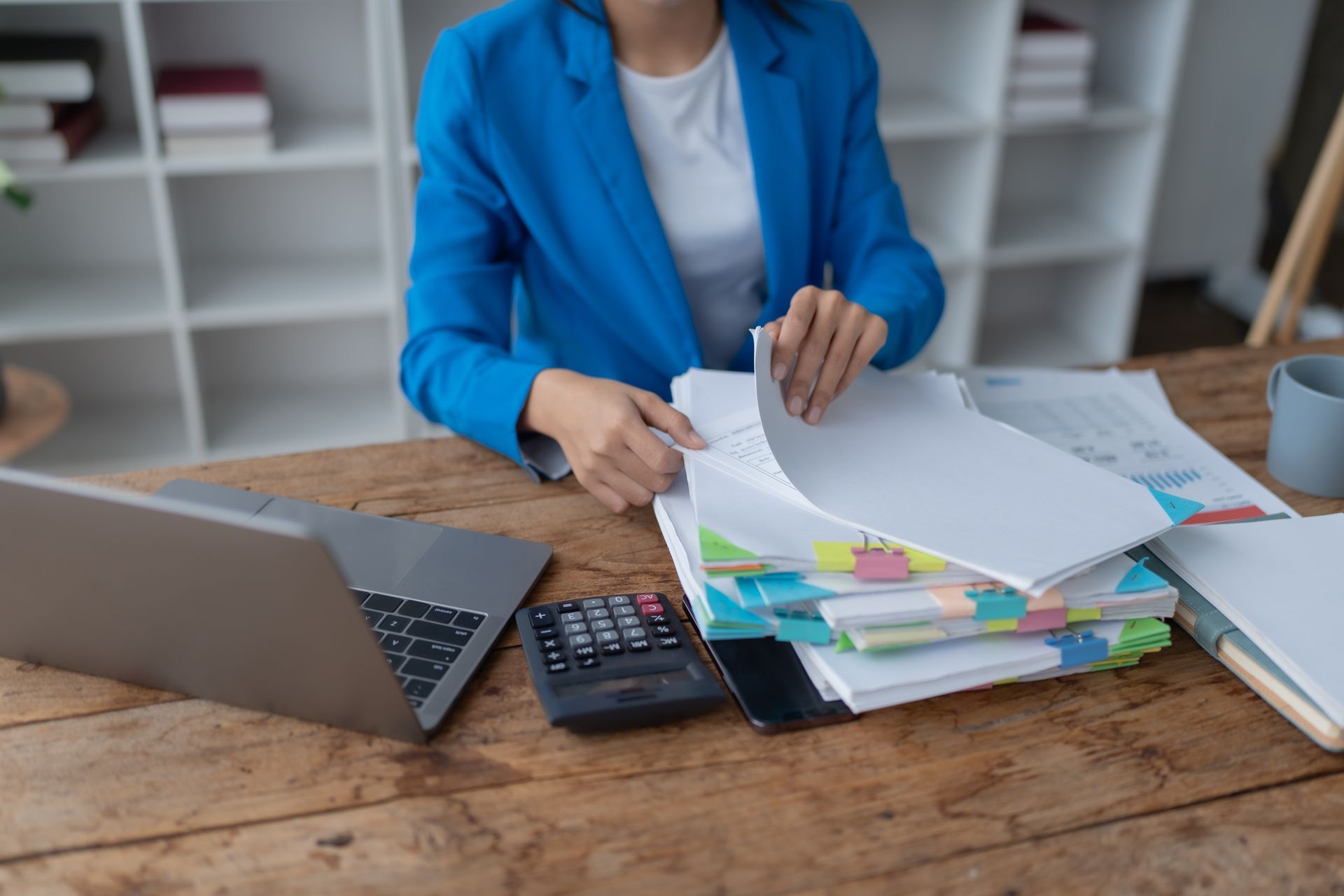 Woman in blue jacket sorts through papers at a desk with a laptop, calculator, and colorful page flags.