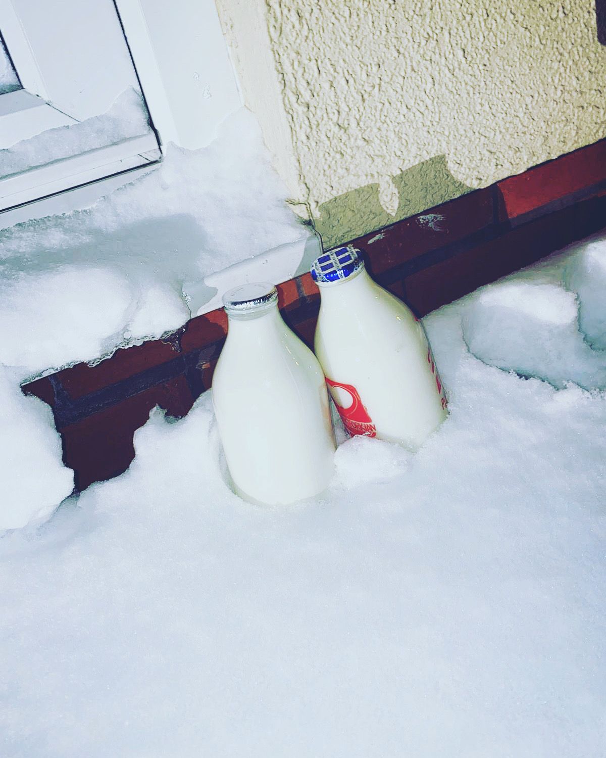 Two glass bottles of milk with foil lids resting in deep snow against the brick wall of a house.