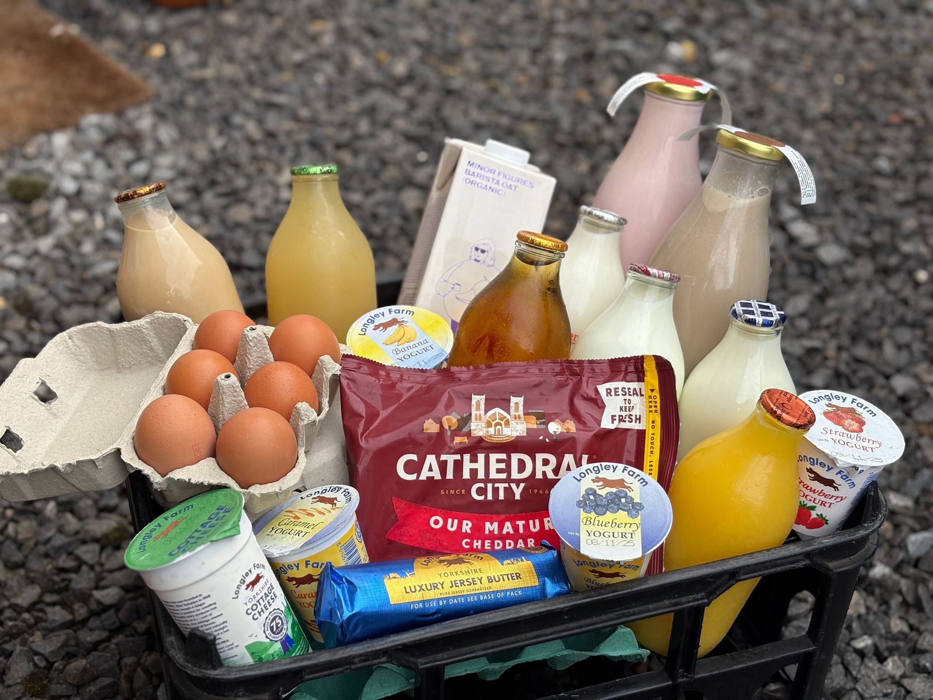 A milk crate filled with grocery items including eggs, cartons of milk, juice, Cathedral City cheese, and yogurt pots.